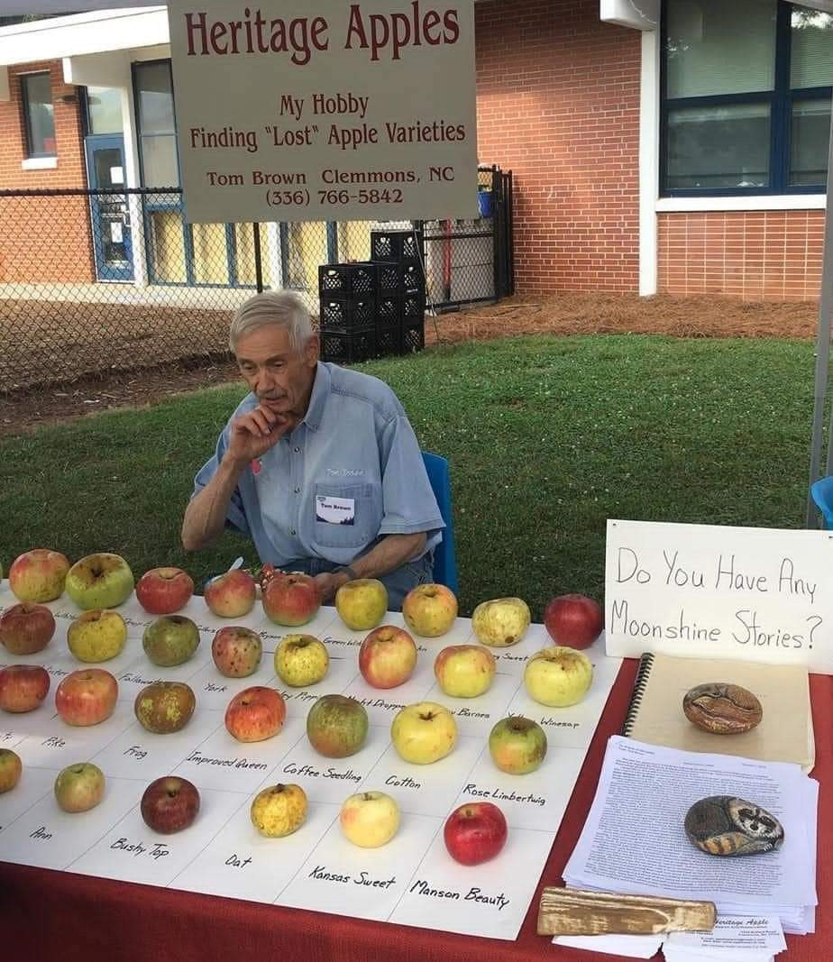 A man in front of his apple collection. A sign says "Heritage Apples. My Hobby Finding "Lost" Apple Varieties." Another sign says "Do you have any moonshine stories?"