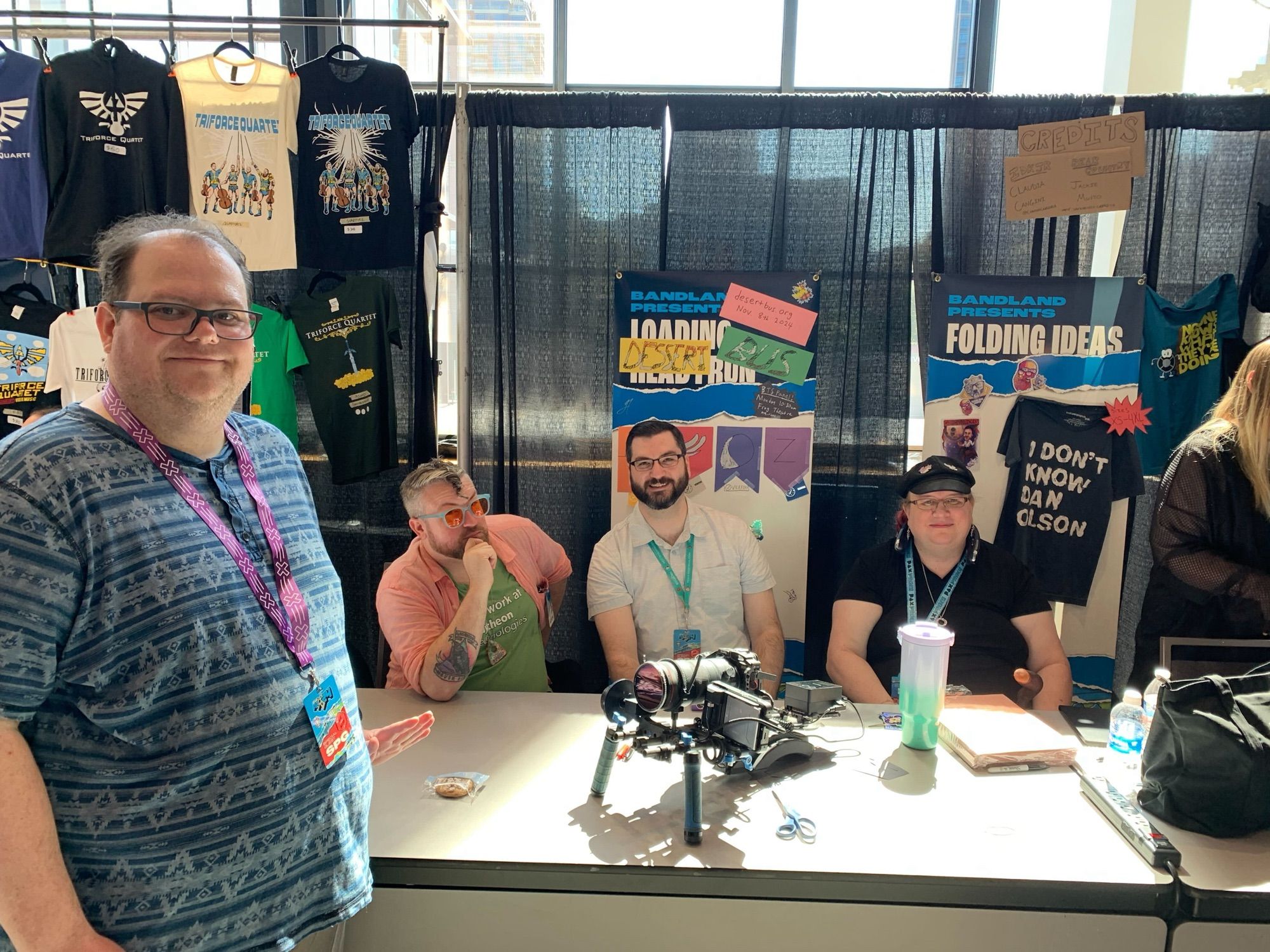 This is a photo of a table at Bandland at pax west 2024. There is a visible loadingreadyrun banner in the background that has been defaced with construction paper to label is as a Desert Bus booth. A bunch of smiling desert bus volunteers are at the table and spiking the camera