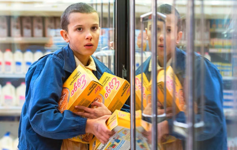 screenshot of Eleven from season one of Stranger Things holding several boxes of frozen waffles in a store