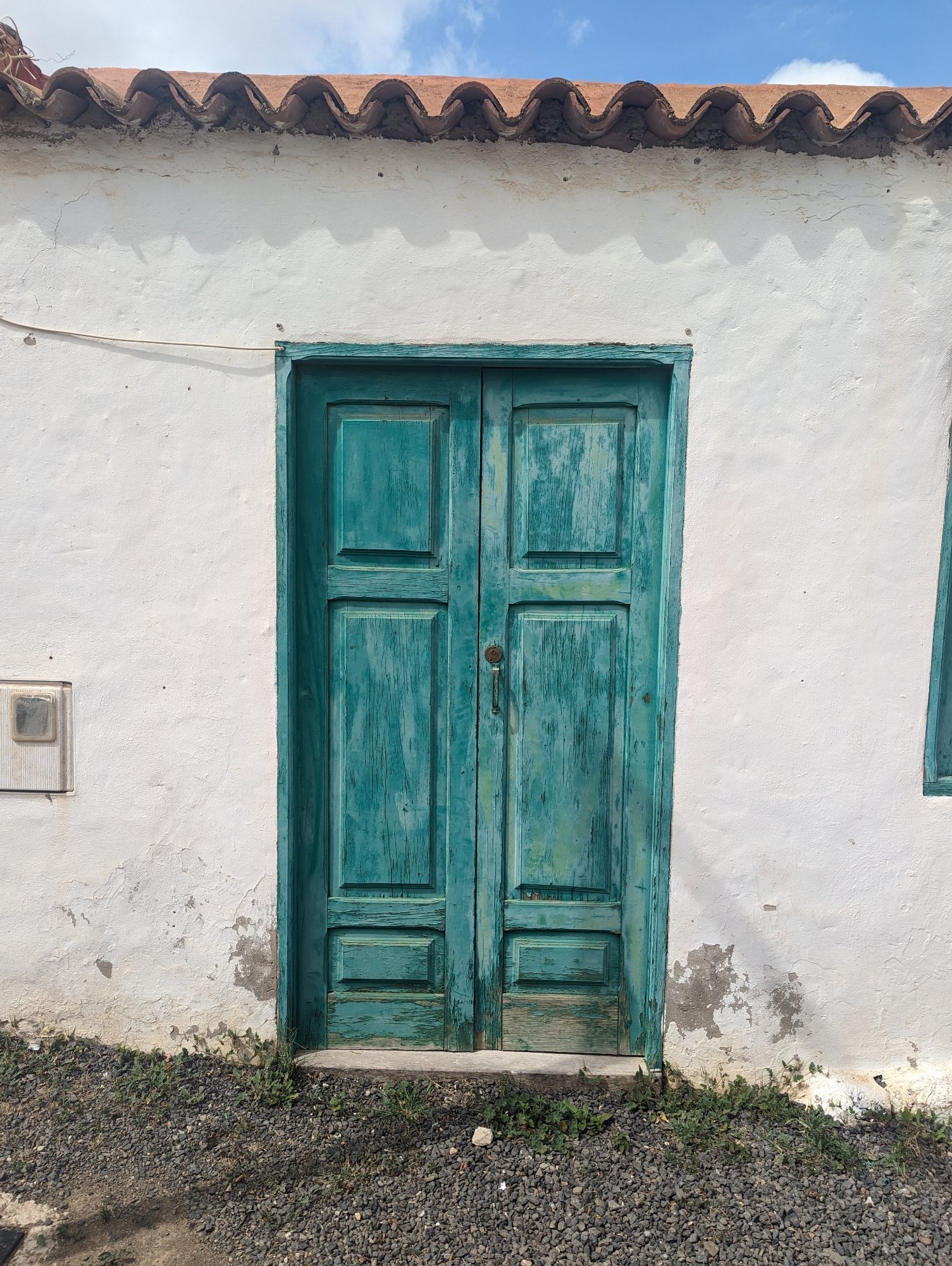 Eine türkisfarbene Holztür, die die besten Jahre schon hinter sich hat, umgeben von einer weiß getünchten Wand. Am oberen Bildrand ist ein Teil des Daches sowie ein Stück blauer Himmel zu sehen. Im Vordergrund ist Kies auf dem Boden