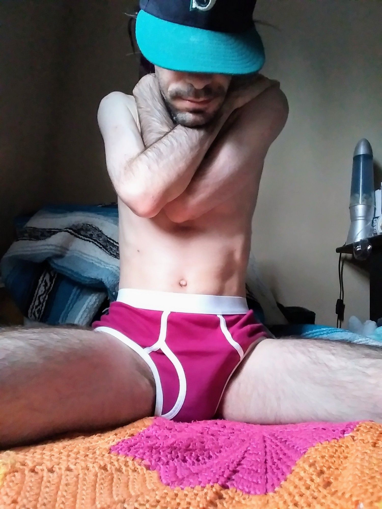 man wearing burgundy briefs, crossing his arms and angling his ballcap to block his face