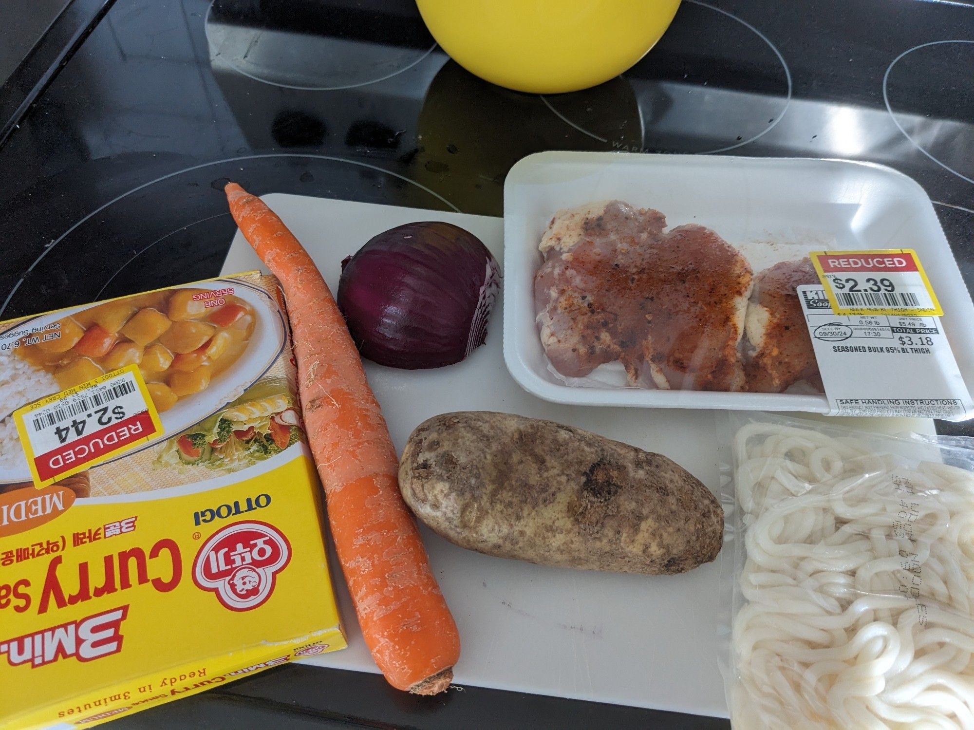 A box of Ottogi curry sauce with a $2.44 discount sticker on it, a carrot, half a red onion, a small potato, a small package of chicken thighs with a $2.39 sticker on it, and a pouch of udon noodles.