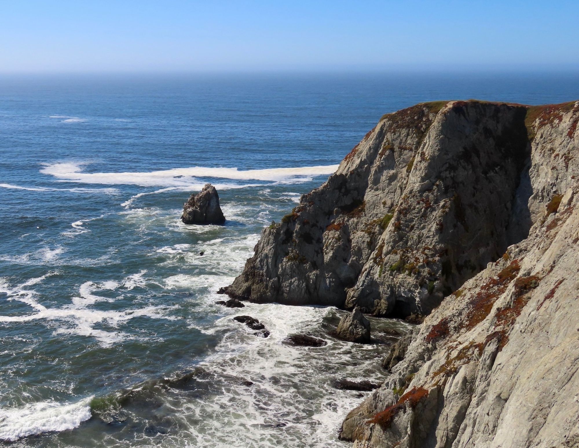 Another long view of granite cliffs to the right and ocean to the left, with wave breaking and white foam on surface.