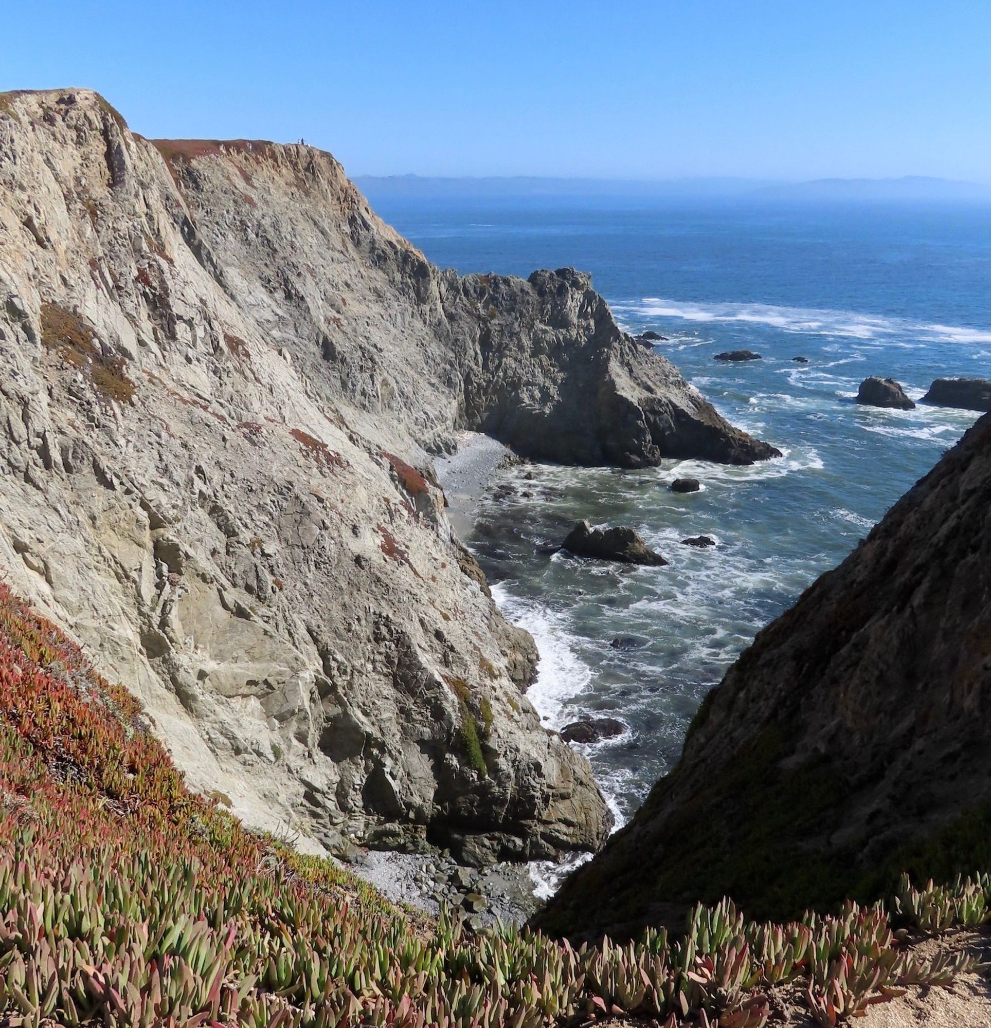 Long shot from about 250 feet above Pacific Ocean with rugged granite cliffs on left and ocean on the right and in distance. Tiny specks of humans are standing at cliff's edge in upper left.