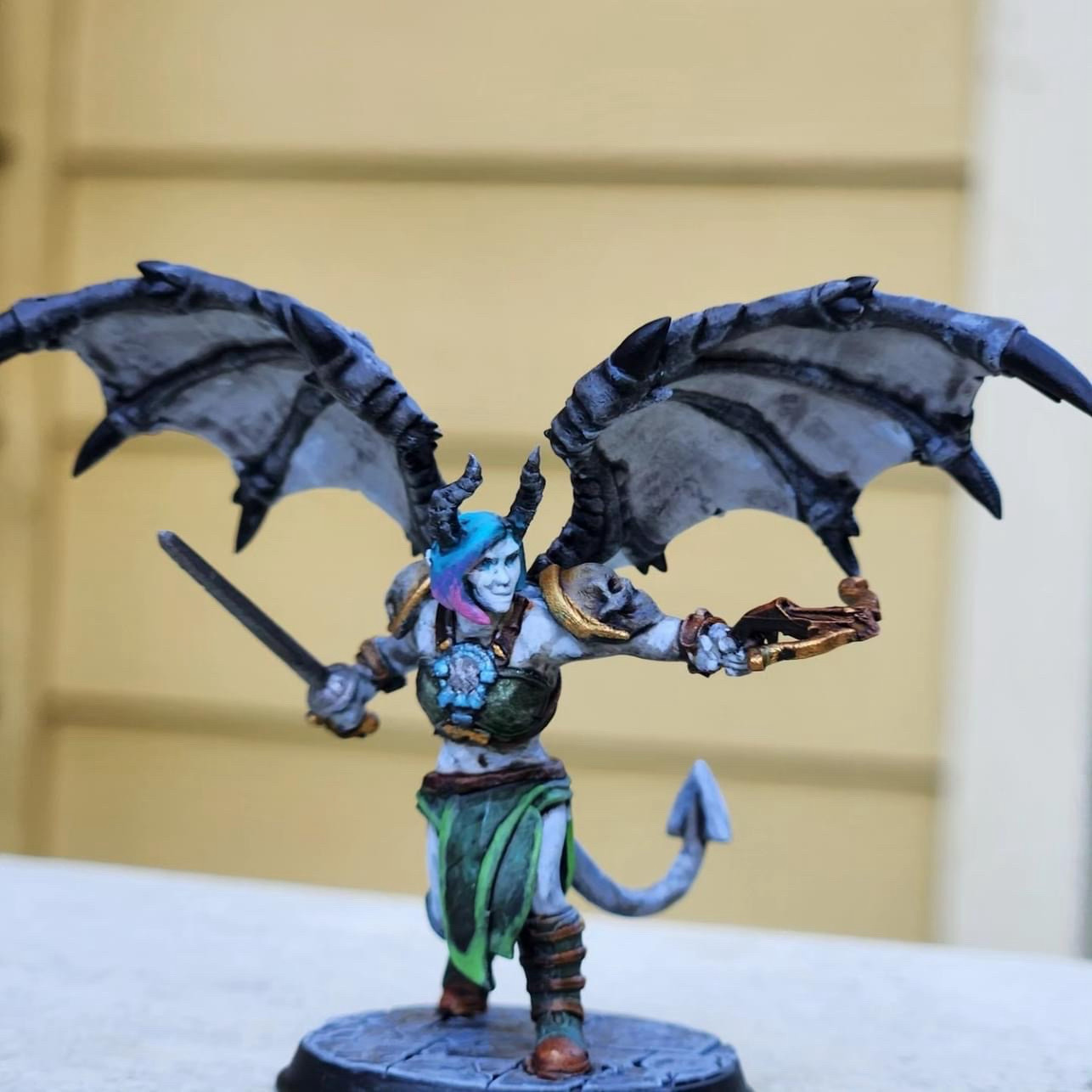 A customer’s photo from a wonderful review of the print and paint work of EPICGMCo. A Grey skinned Winged Tiefling with black horns and in hunter green garments sports a gradient blue-to-pink hair while they hold a Silver sword and a Golden Hand Crossbow in an action pose. The colors are vibrant.