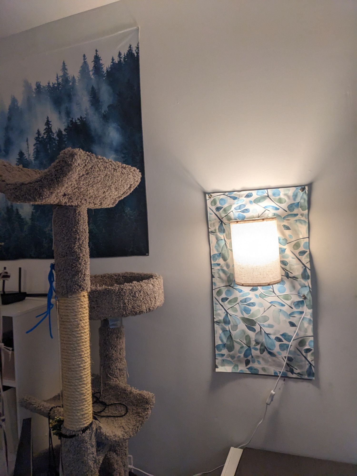 A square of blue toned leafy fabric hangs on a white wall to conceal a utility box. It is held up with magnets. There is a neutral colored lamp hanging over the square, also attached with magnets. There's a cat tree on the left.