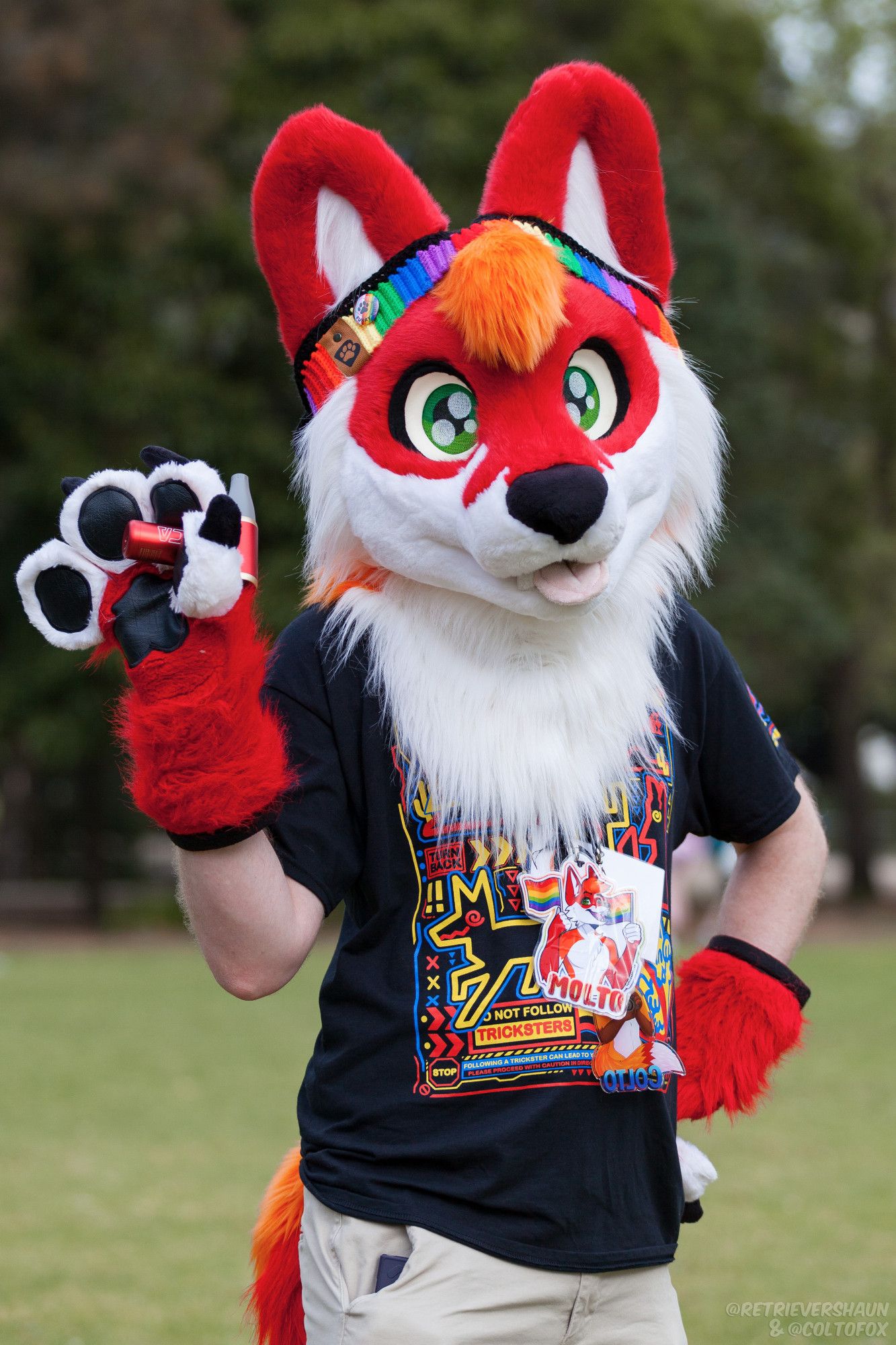 Molto Fox (red, white, black and orange fursuiter) standing in a park holding a Kica fan, in partial fursuit with visible arms.