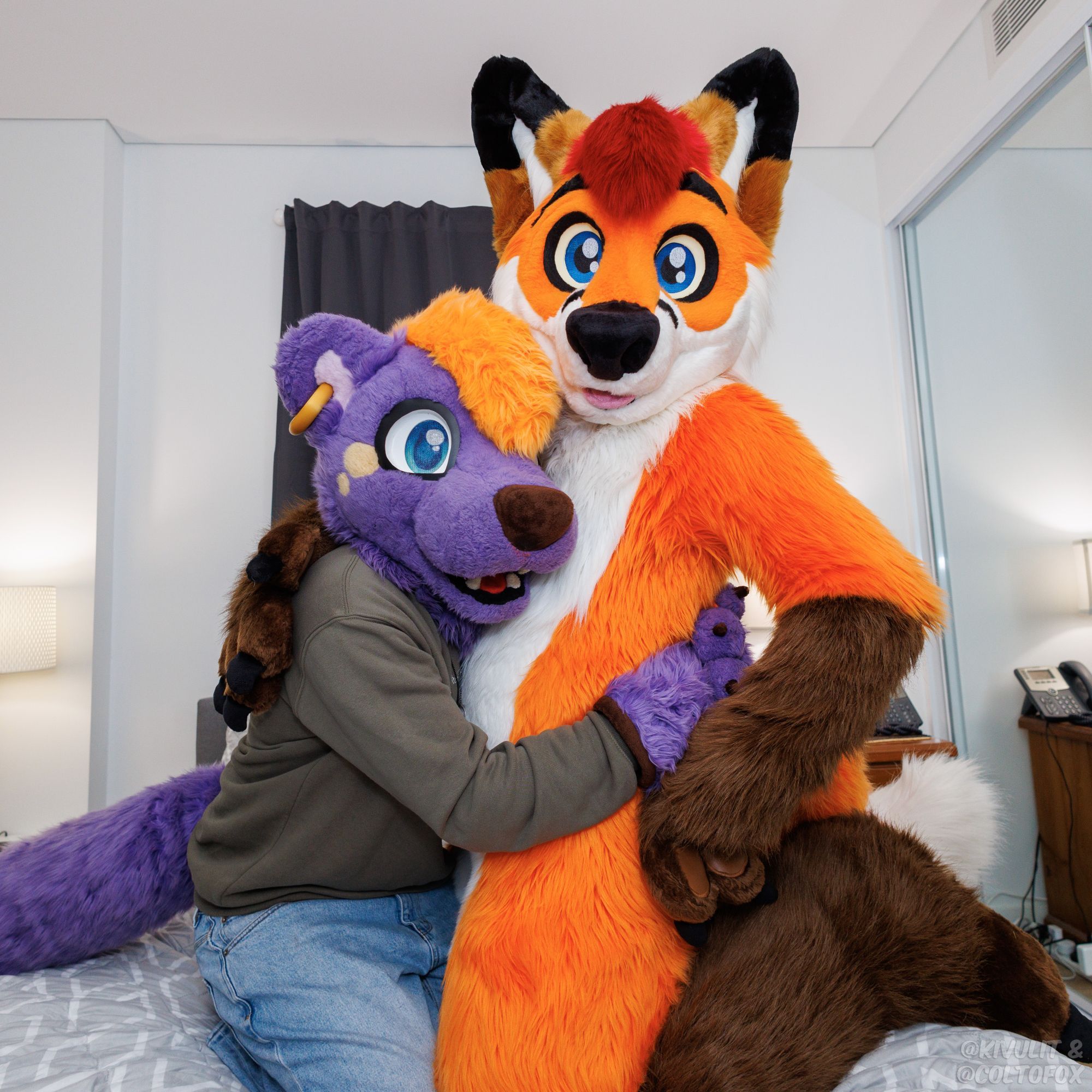 A tall orange fox fursuiter being hugged by a shorter purple otter fursuiter on a bed.