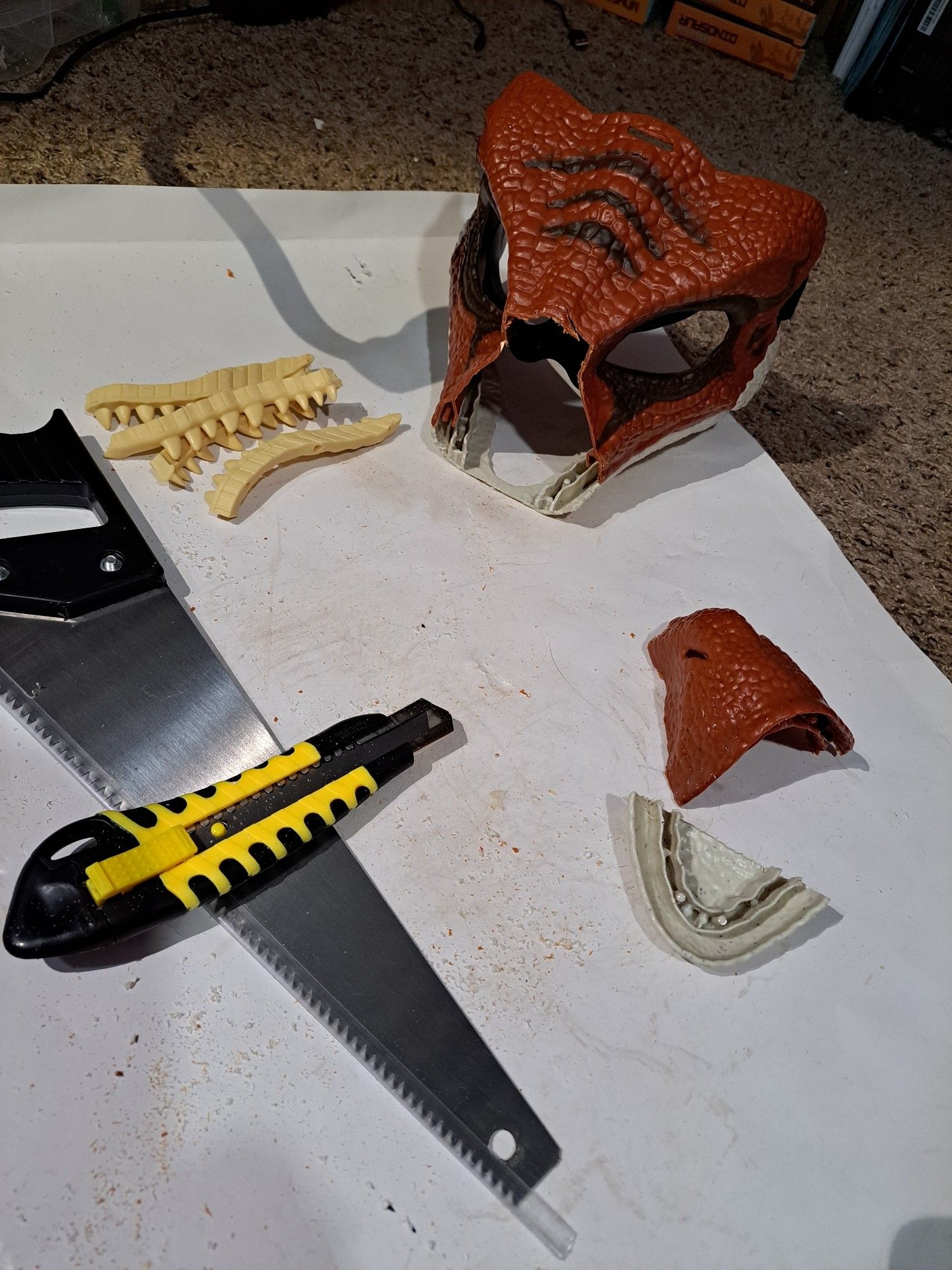 A red dino mask, the front of which has been cut off. The discarded pieces sit to the right. To the left are the removed teeth inserts, and below those sit a cheap handsaw and a box cutter with a yellow and black case. All of this sits on a white tray littered with plastic shavings from the cutting process.