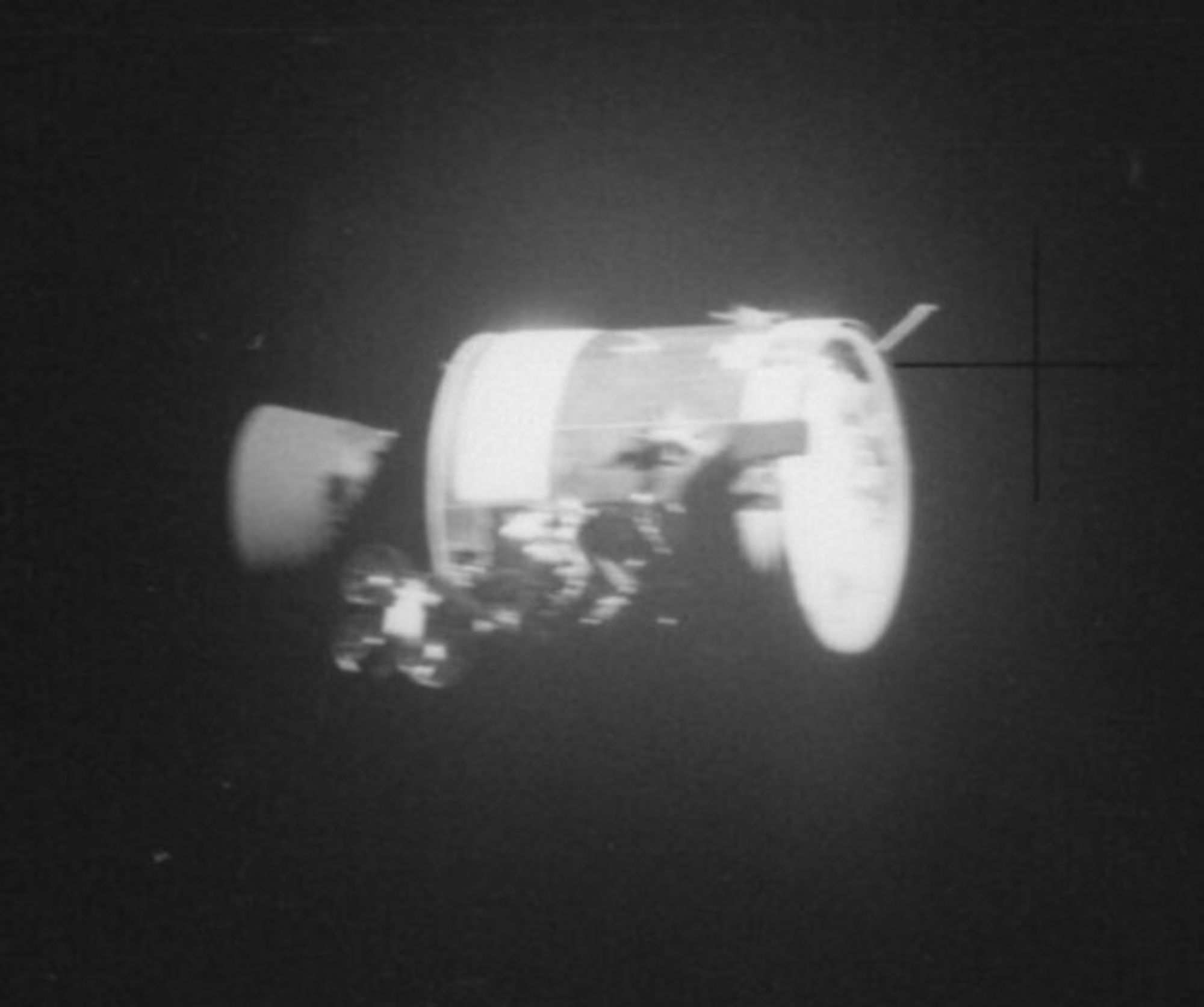 A black and white photo of the Apollo 13 Service Module taken through the window of Lunar Module Aquarius. The service module is far into the dark space and the damage from the explosion who aborted the moon landing mission is visible on its hull.