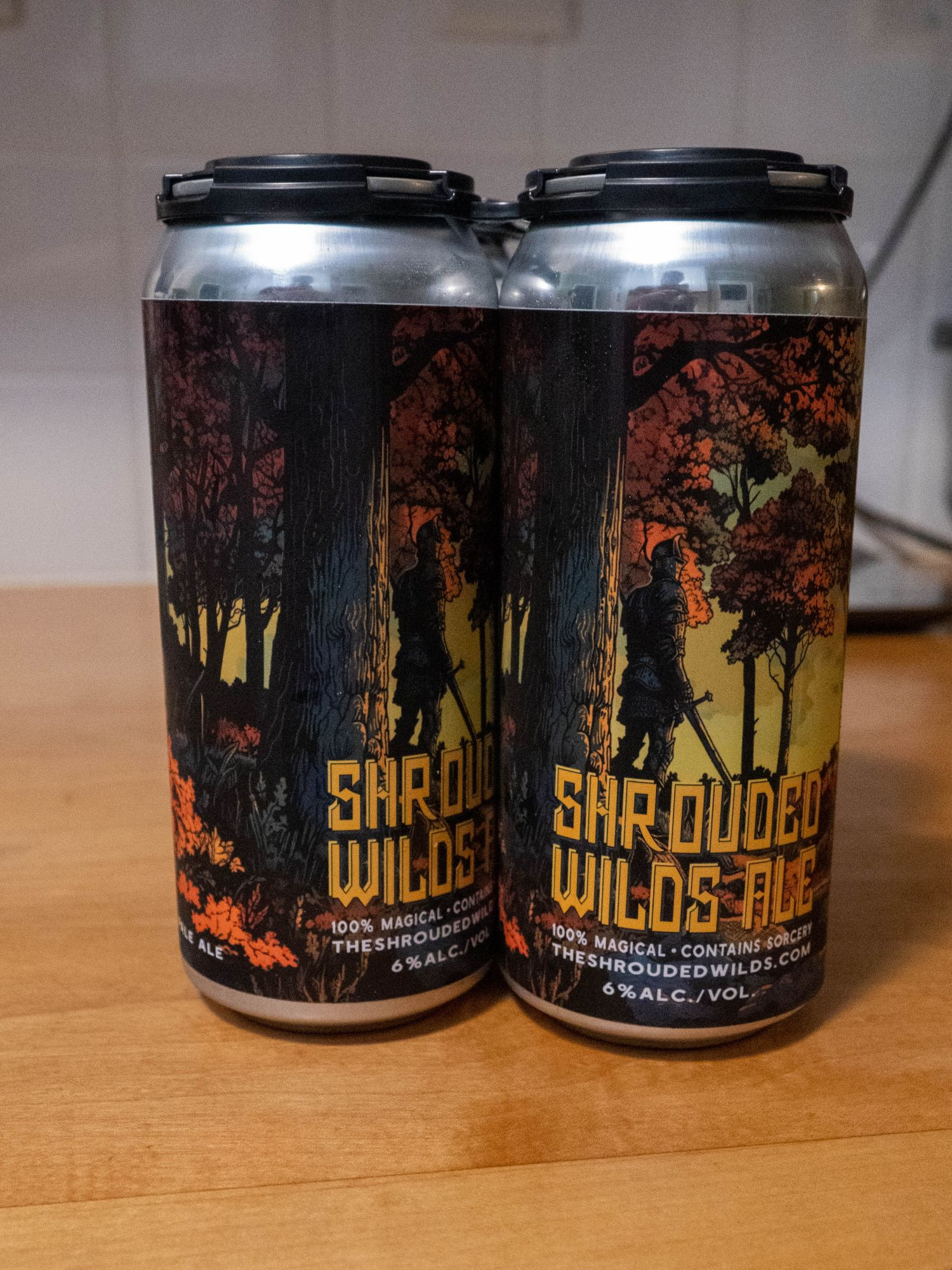 A 4-pack of beer cans showing an adventurer facing a forest and labelled "Shrouded Wilds Ale"