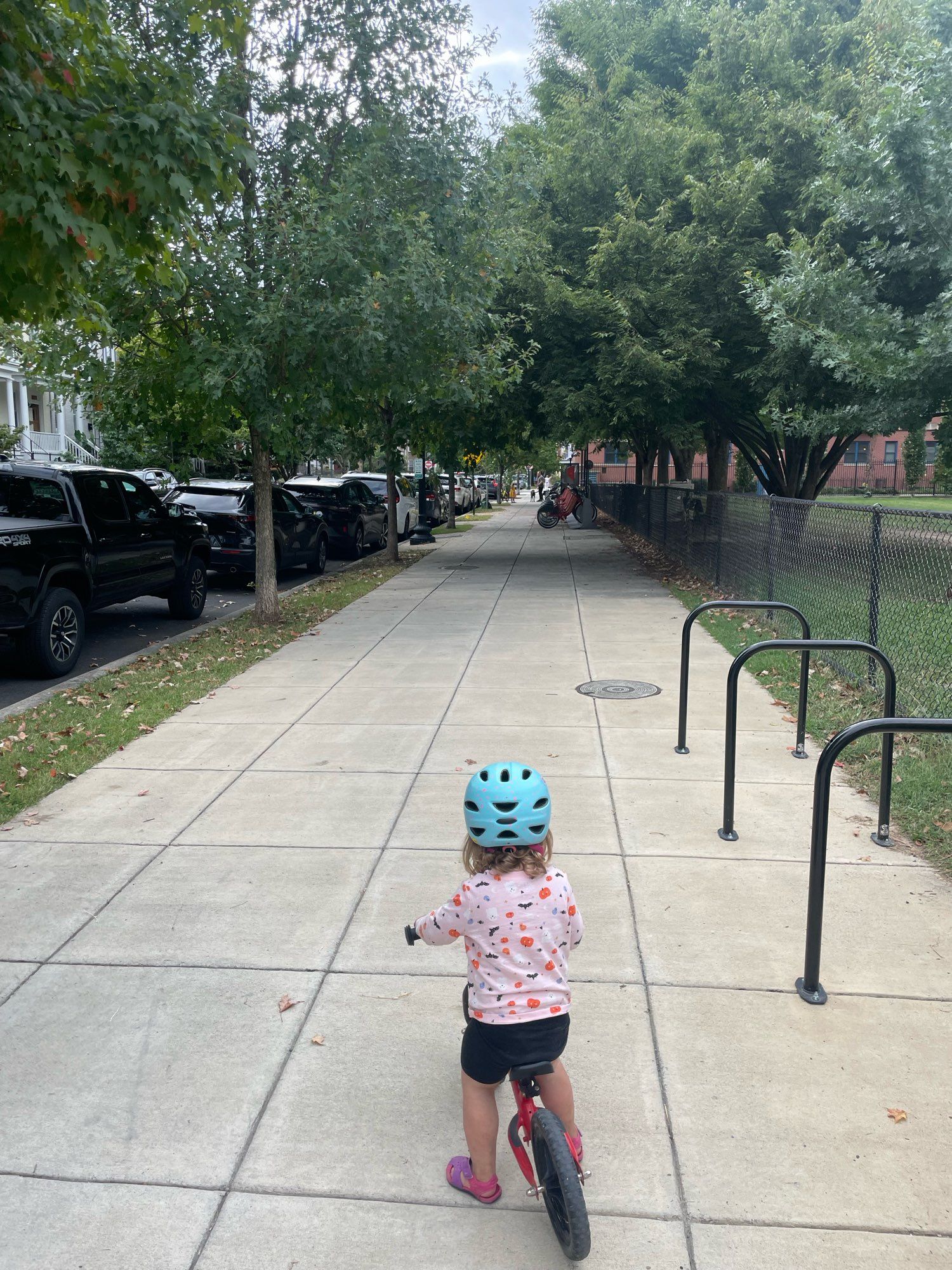 Girl facing away on balance bike near bike racks and a wide sidewalk