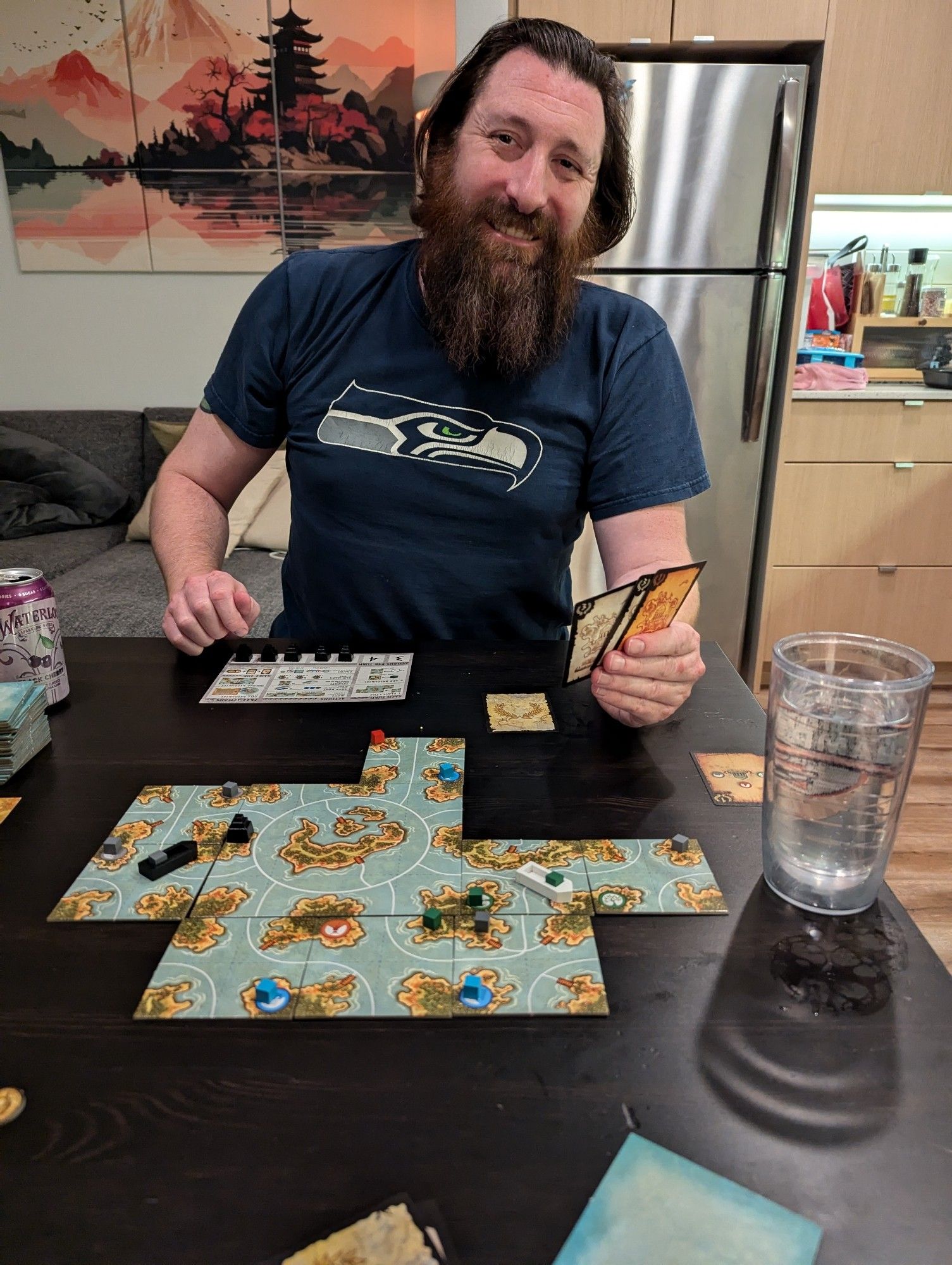 A person at a table with a board game set up, they smile at the camera and have a Seattle Seahawks T shirt on.