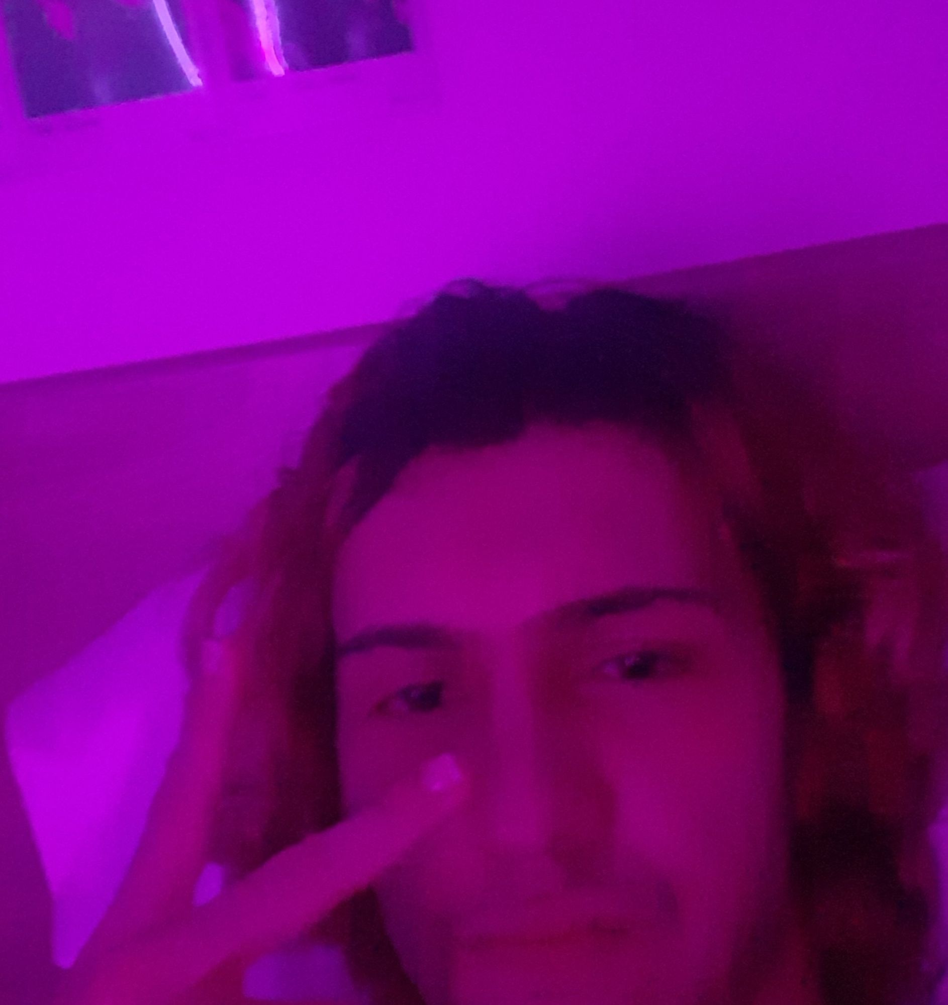 White guy with overgrown roots laying down on bed posing for a selfie and doing the peace sign. There's a pink light iluminating the whole shot