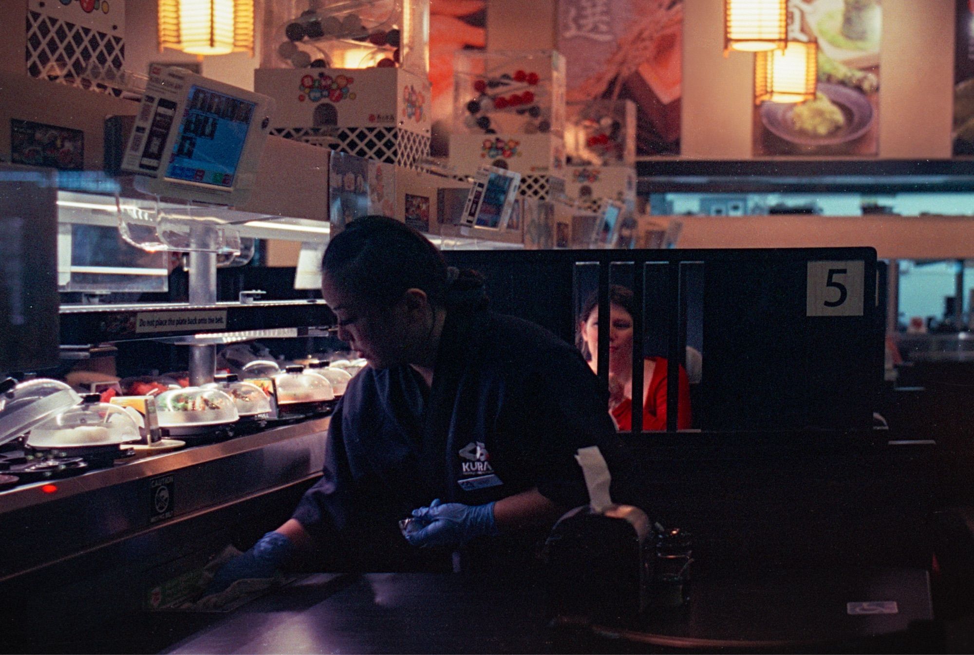 Woman cleans a table at a sushi restaurant while another woman converses in the next booth behind her