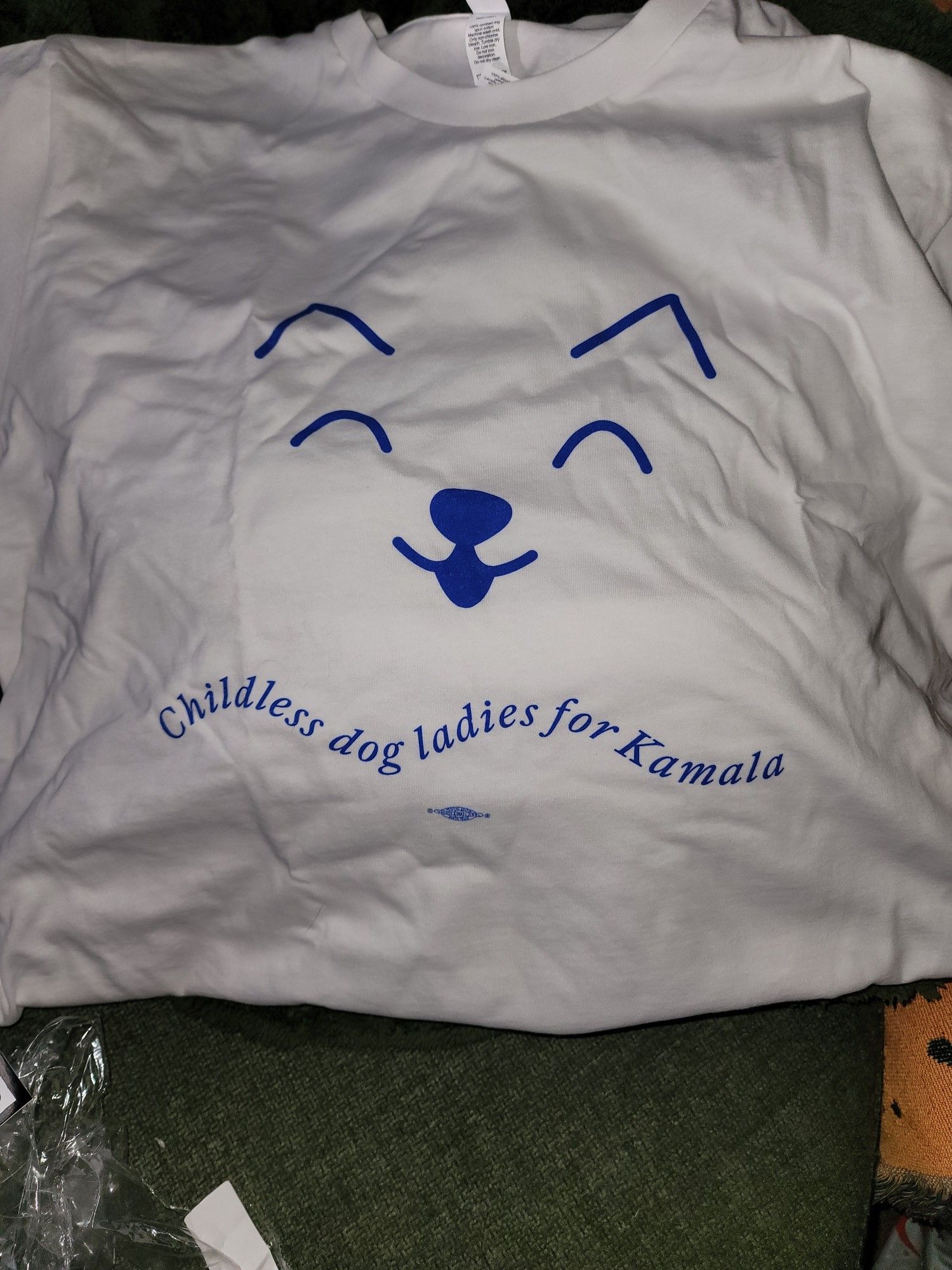 White tee w/ blue graphics of a dog nose, eyes & mouth. Text underneath reads "Childless dog ladies for Kamala".