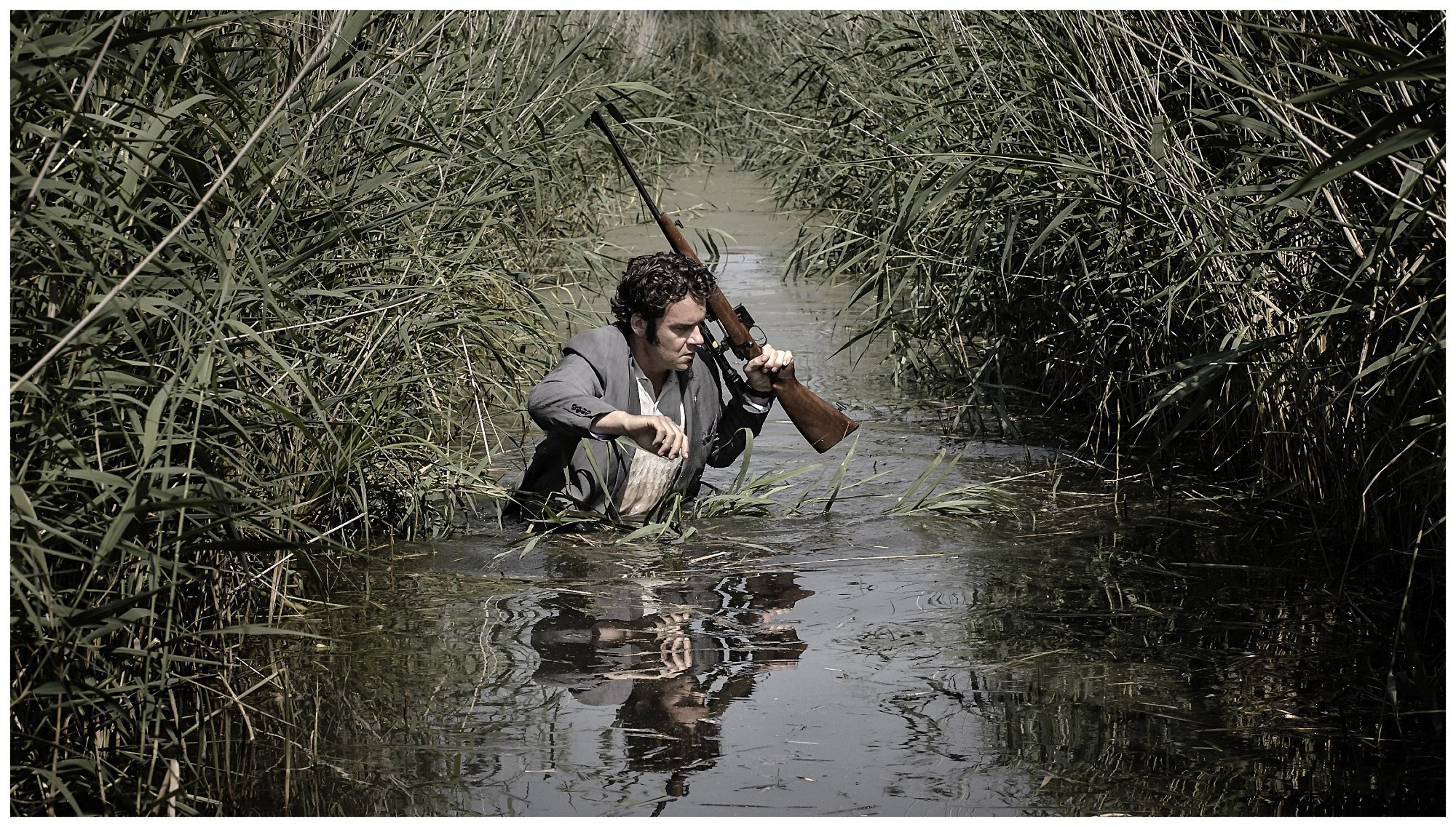 Tom Dewispelaere is seen wading through a swamp with water up to his midriff while holding a rifle, from Alex Van Warmerdam's Schneider Vs. Bax (2015).