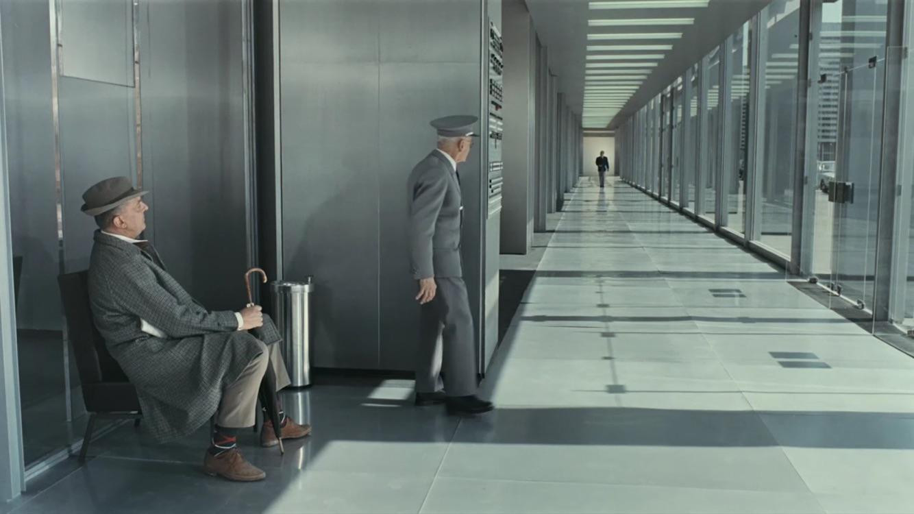 Jacques Tati, dressed as his M. Hulot character, sits holding an umbrella in a long, modern hallway. An older attendant stands nearby, peering down the hall at a distant, approaching figure. From Tati's Playtime (1967).