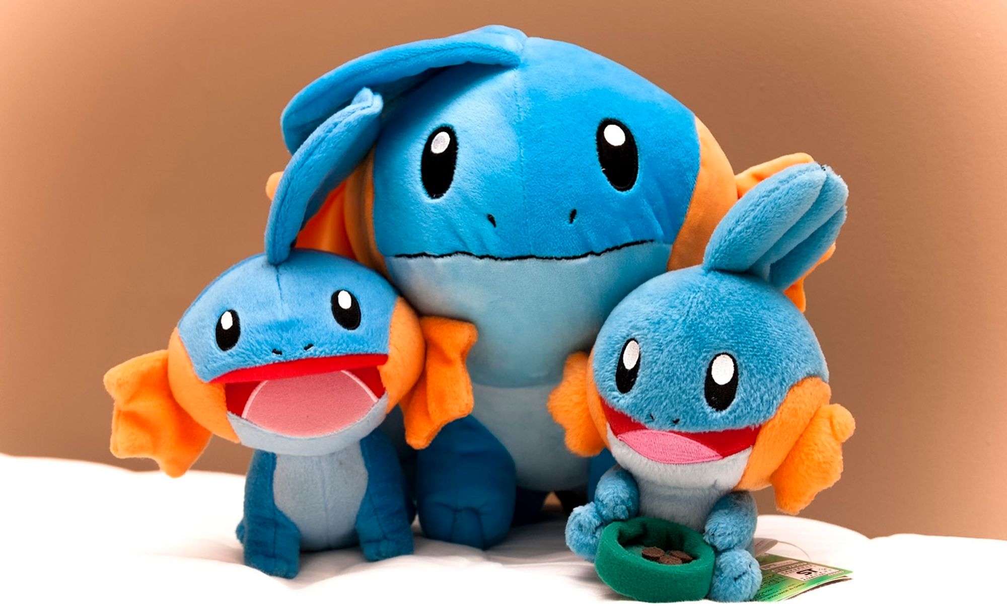 A photo of three mudkip plushies, with one larger one in the middle and two smaller plush by its side