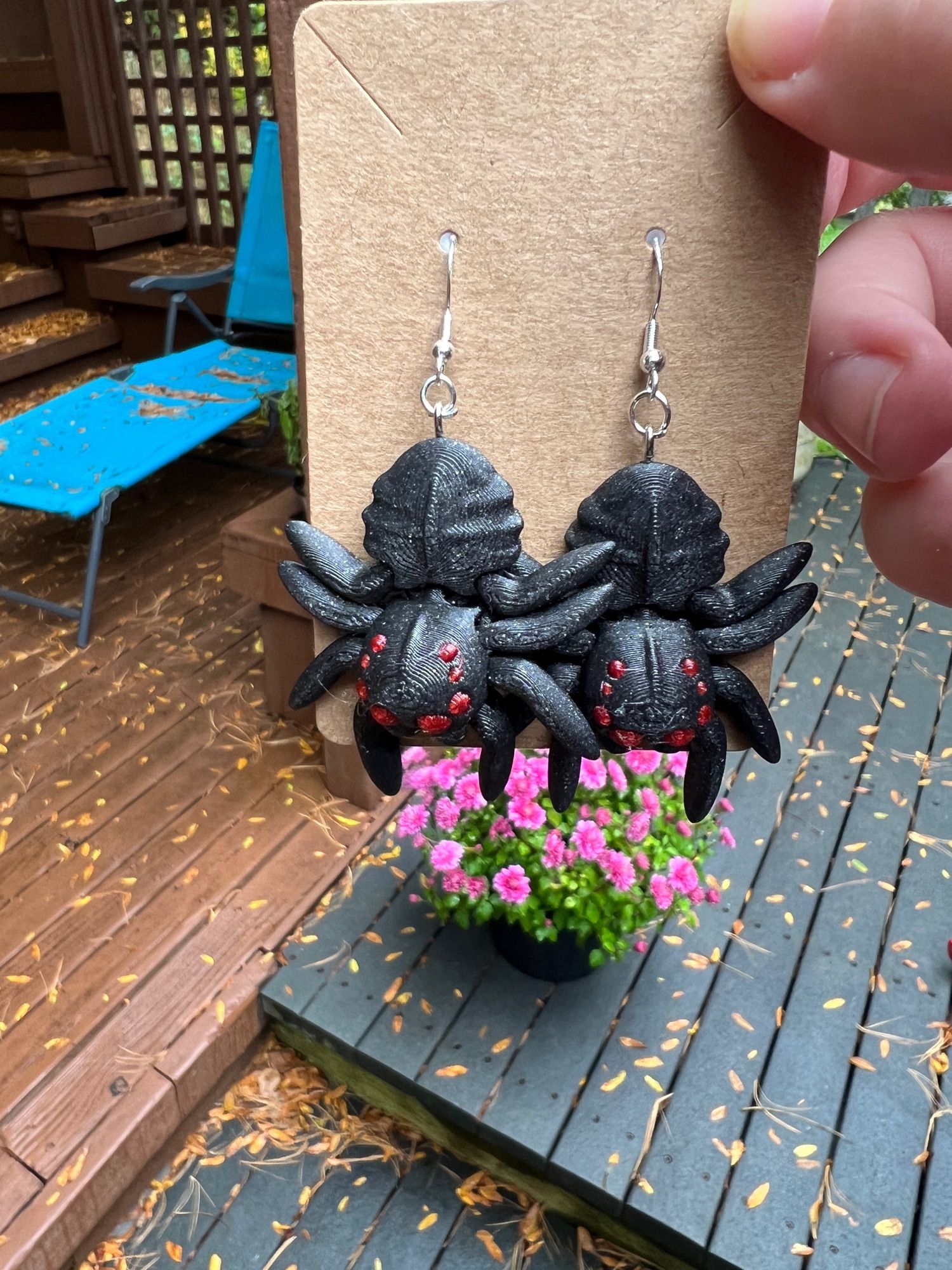 Sparkly black and red spider earrings on a cardboard backer with a pink mum in the background.