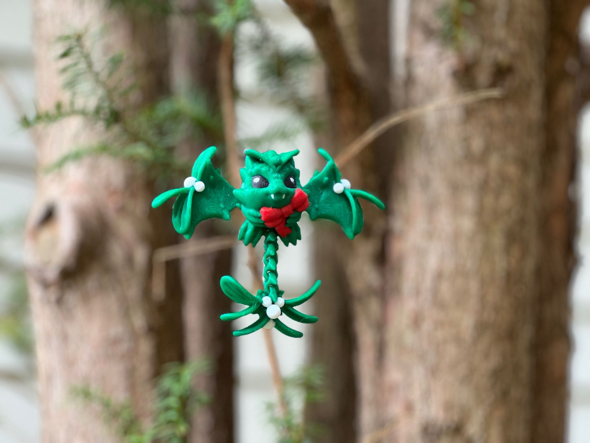 Baby mistletoe dragon 3D printed in green holding a red bow in front of some trees