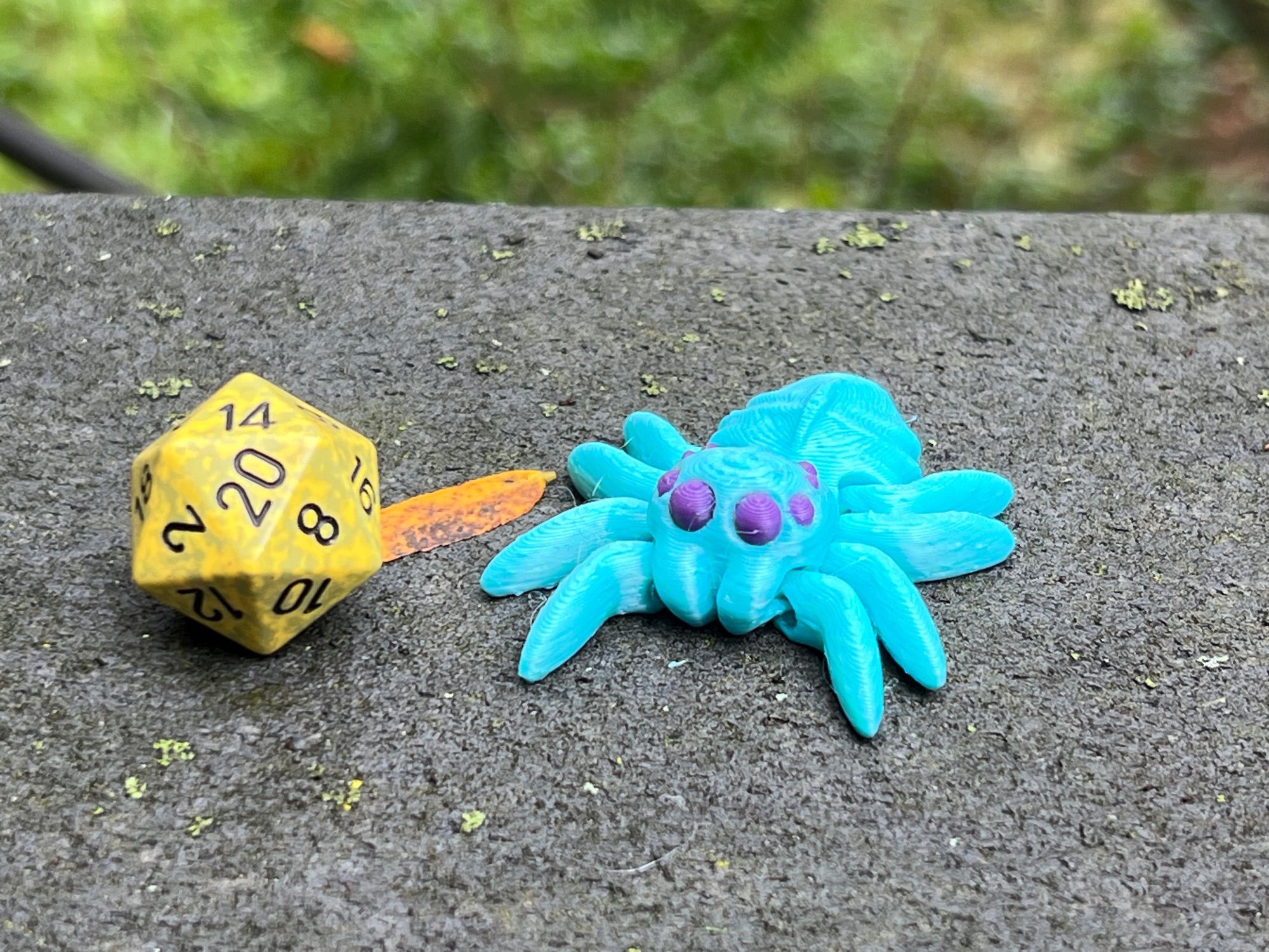 A teal spider with purple eyes next to a greenish d20