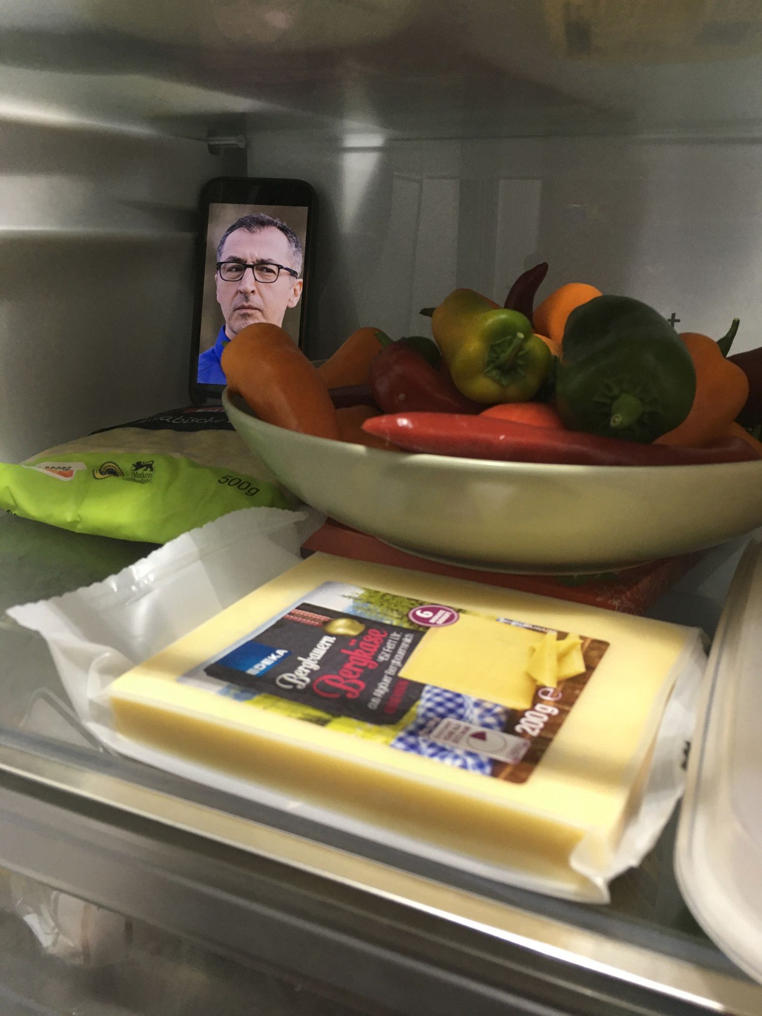 Handy mit Bild von Cem Özedmir der im Kühlschrank auf eine Schale mit Paprika schaut. Davor liegt eien Packung Käse.