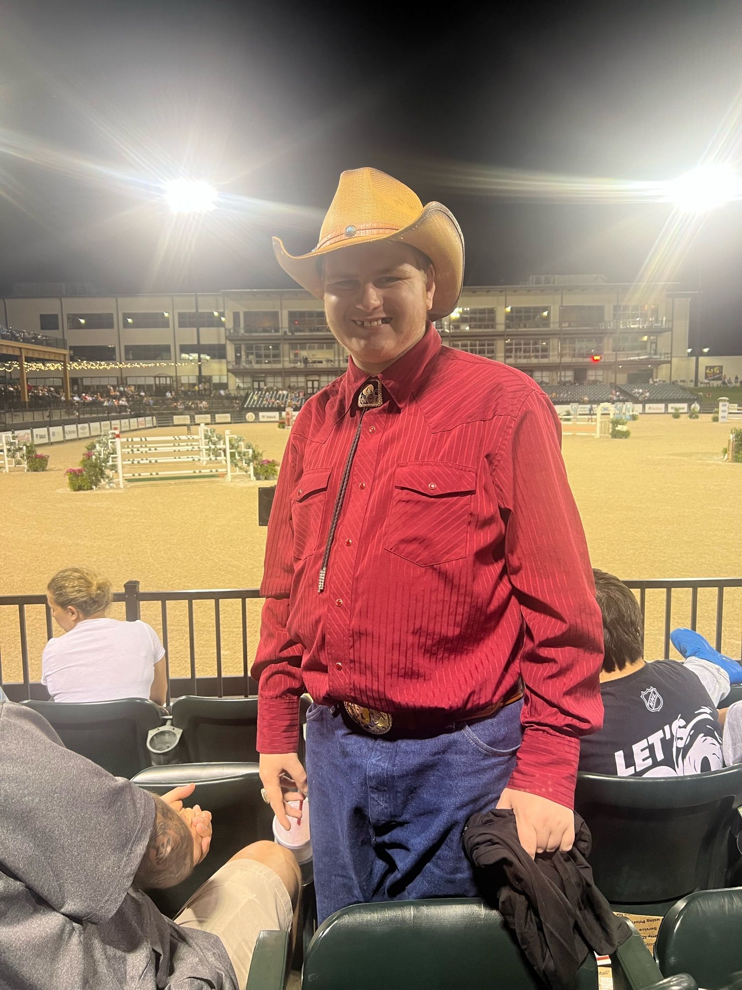 Photo of a Special Olympics North Carolina athlete (Jacob Huffman) standing up at Saturday Night Lights at the Tryon International Equestrian Center. They are wearing denim cowboy-cut jeans, a red western shirt, a bolo tie, and a straw (tan/brown) cowboy hat