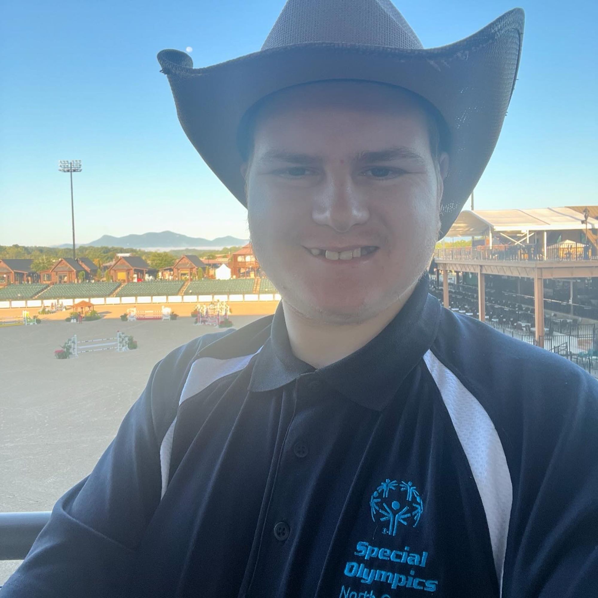 Photo of a Special Olympics North Carolina athlete (Jacob Huffman) wearing a black polo with white stripes on the shoulders with the words “Special Olympics North Carolina” on the chest. In the background is the Tryon International Equestrian Center and Resort