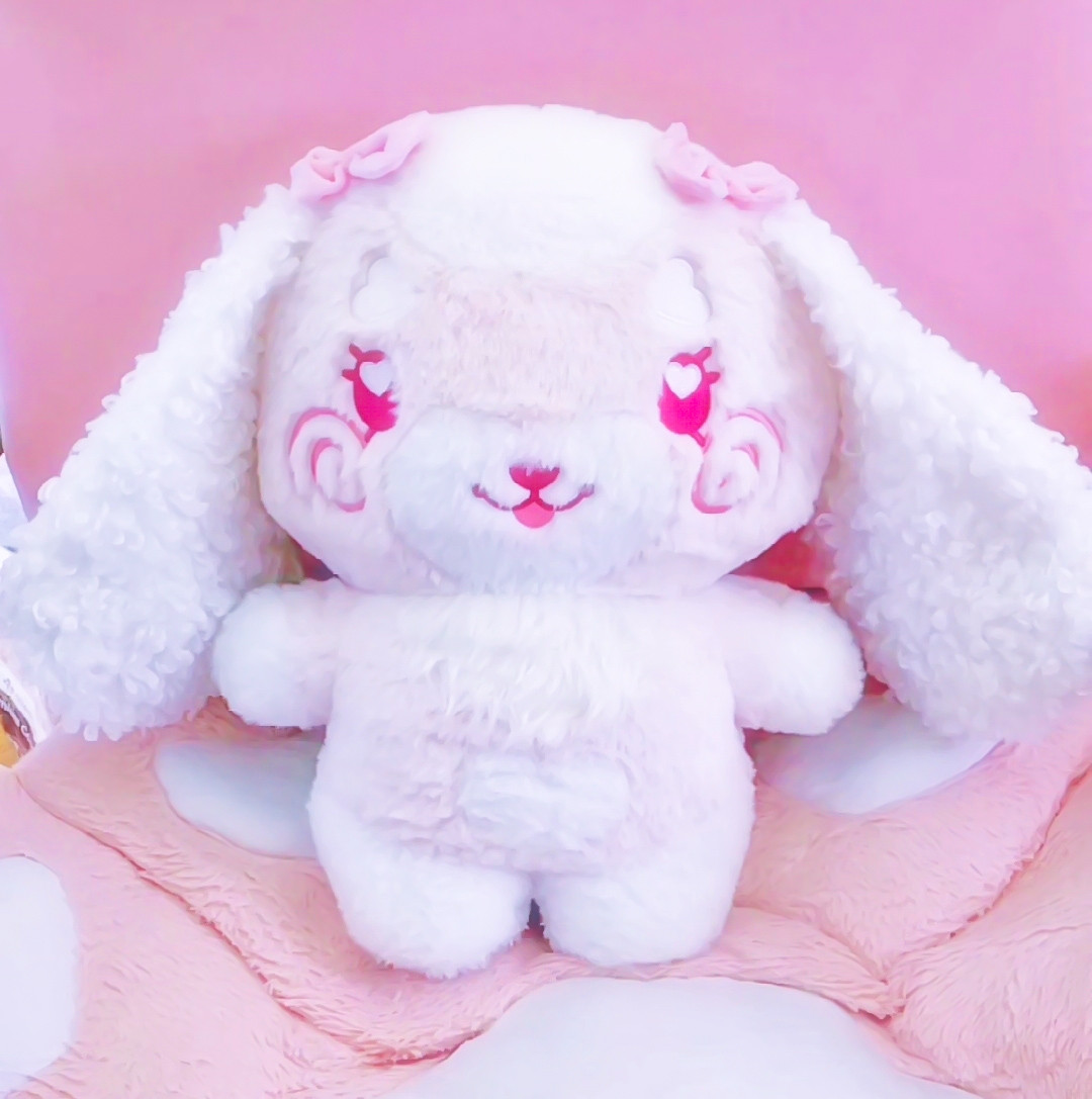 A stylized bipedal chibi plush of a mostly pink and white lop bunny character plushie with swirly blush cheeks, pink eyes, fluffy white ears with two pink bows above each of her floppy long ears. She's seated on a pink chair and large pink cat paw cushion.