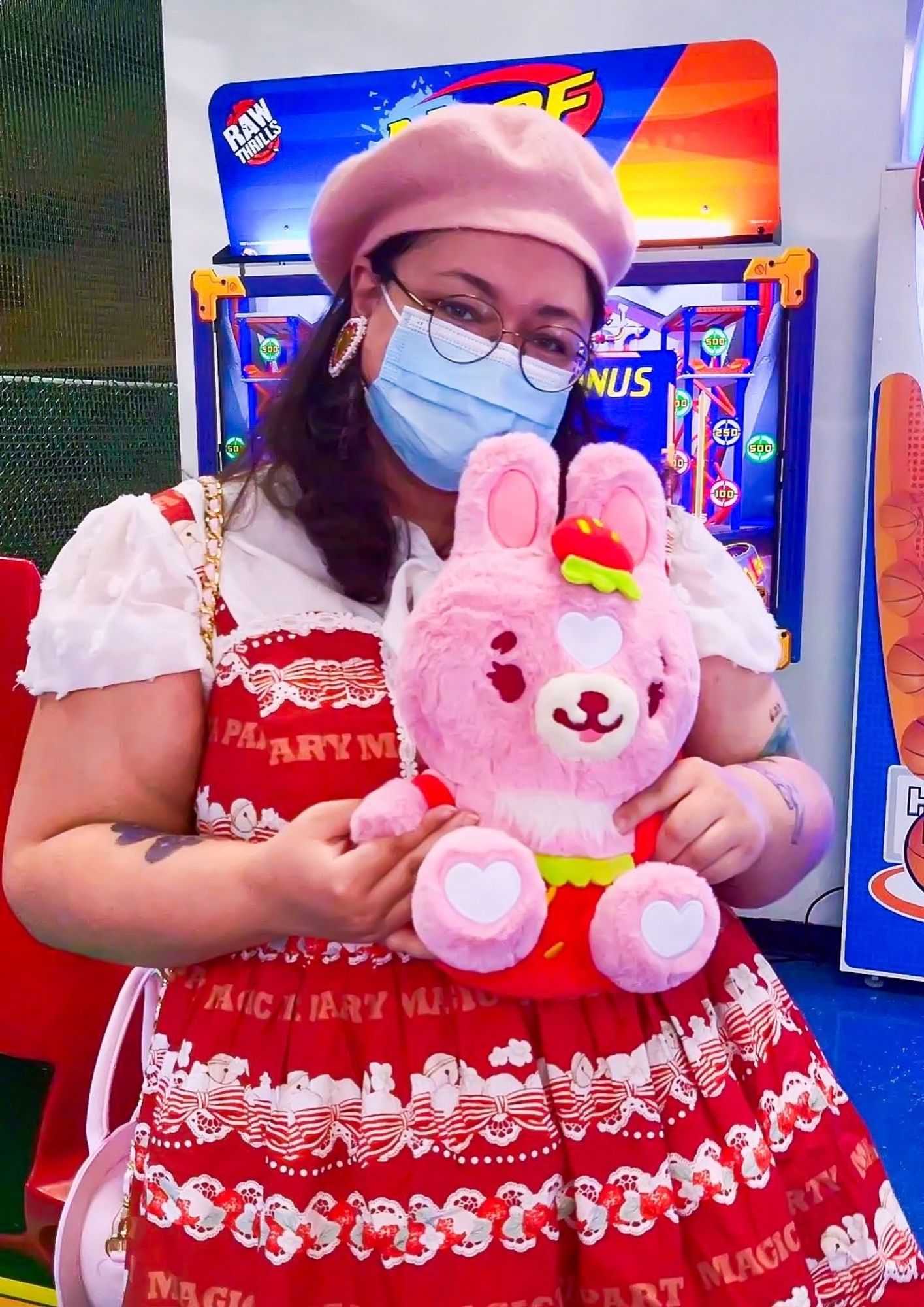 A person in a red puffy dress at an arcade holding a stylized pink bunny plush. The plushie is wearing strawberry overalls and a matching strawberry hat.