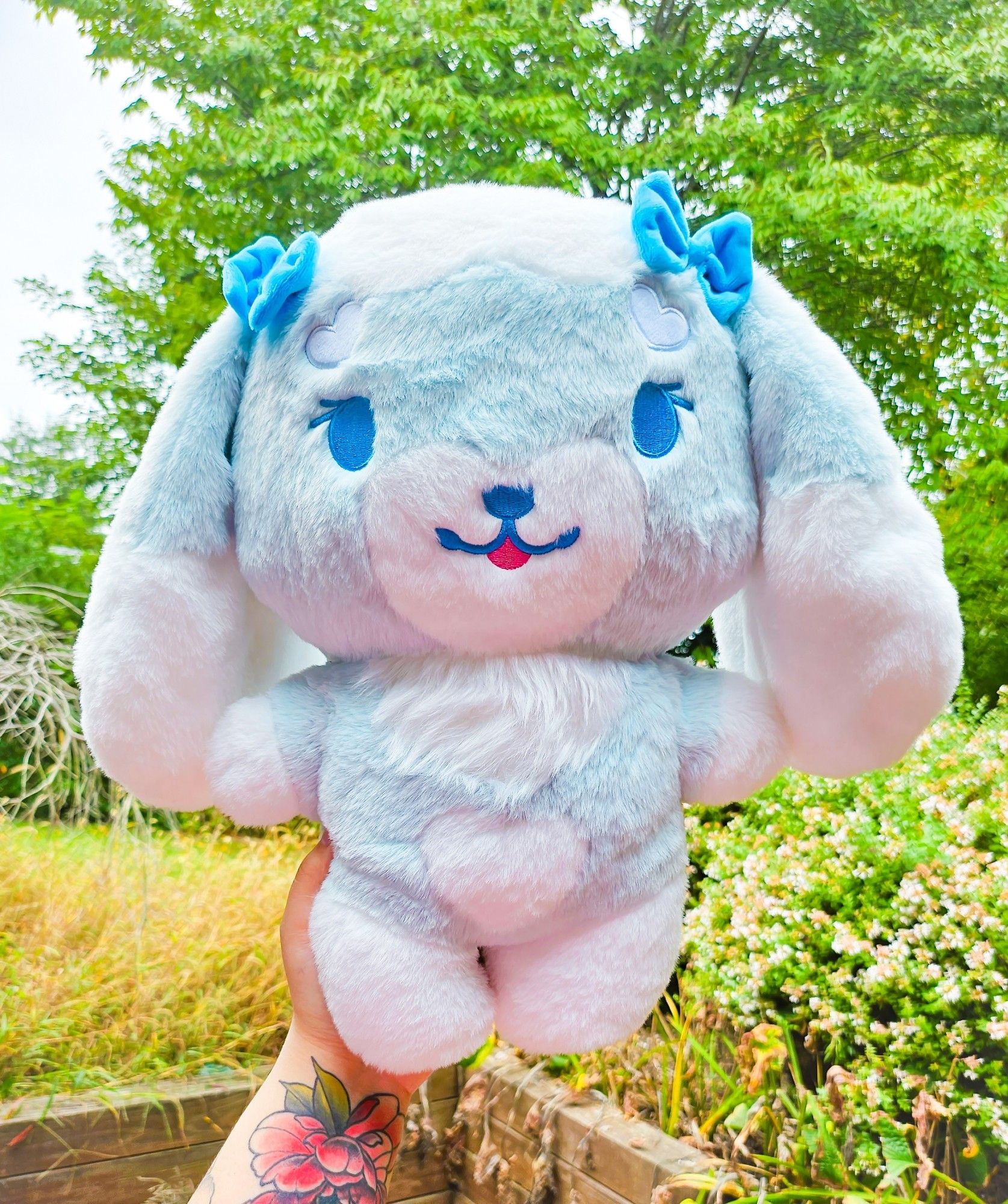 A large, stylized, bipedal light
blue and white lop-bunny character
plushie with two blue bows on the top of her ears and embroidered blue eyes and facial features.