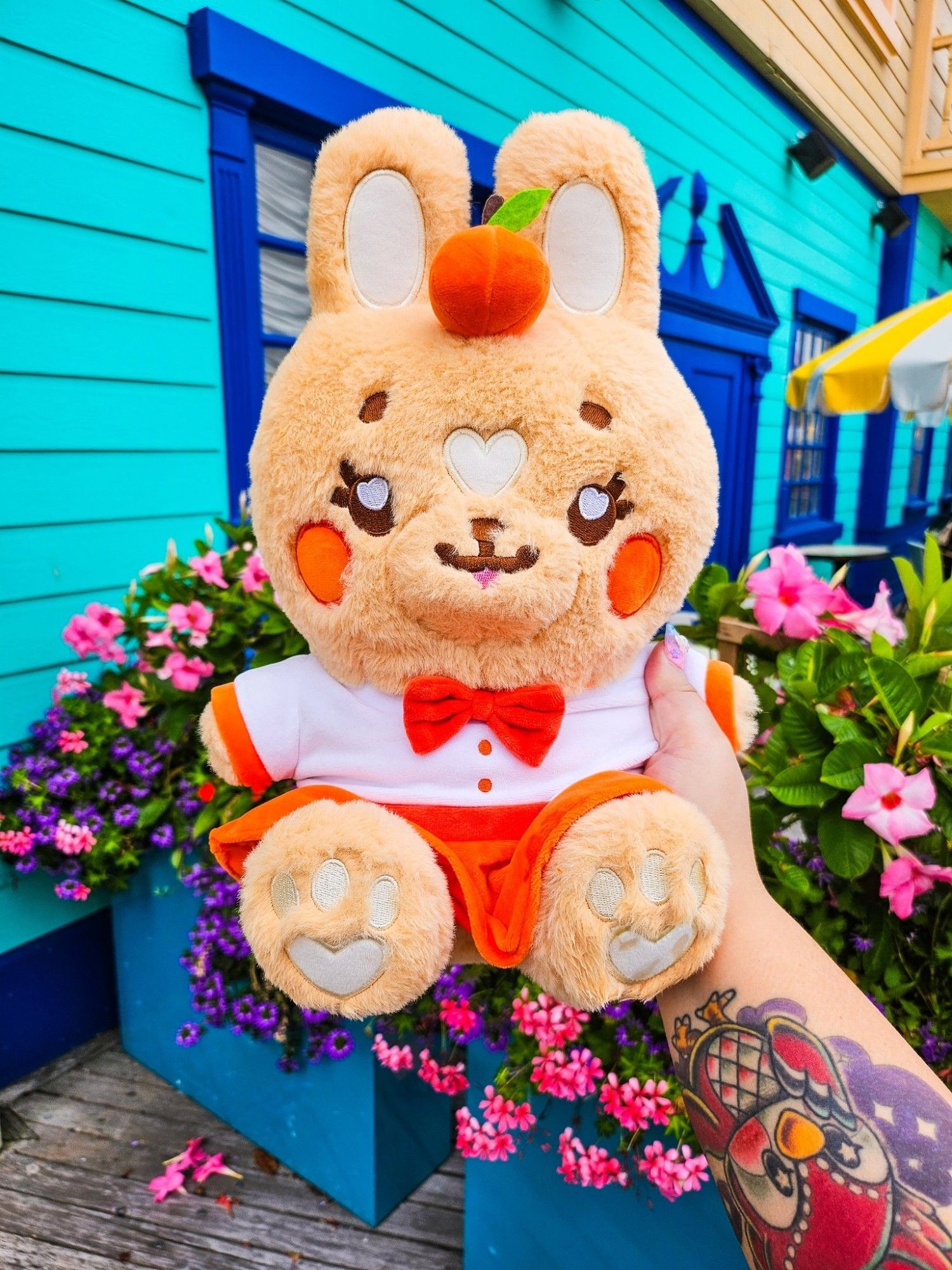 An orange rabbit plushie in front of a
bright blue house with colorful pink
and purple flowers as well. The plush
wears a white blouse with an orange
skirt and tangerine fruit hat on her
head.