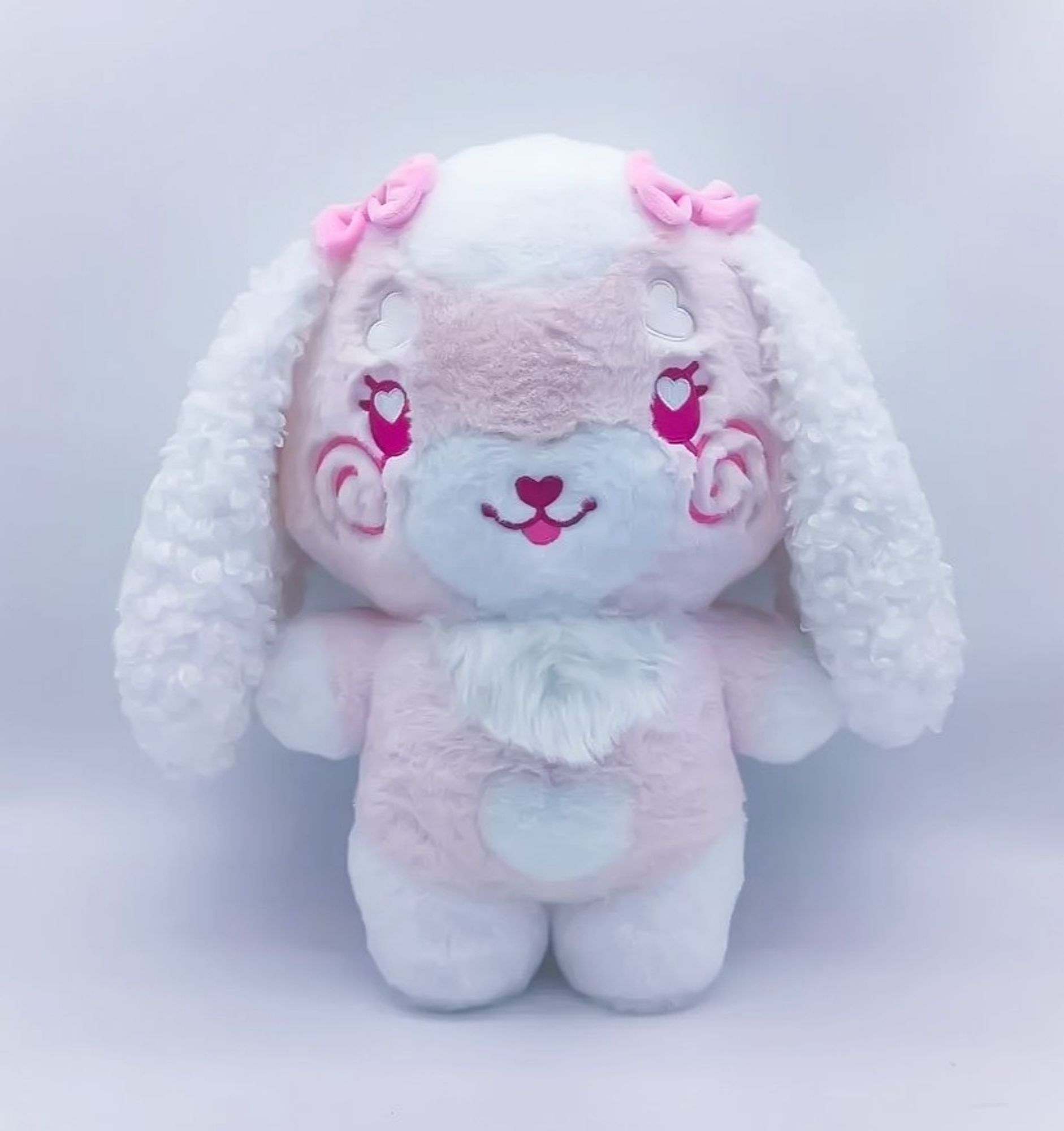 A stylized bipedal fluffy mostly pink
with various white accents lop-bunny
character plushie with pink eyes with
white hearts in them, and spiral pink
blush on her cheeks. Her ears are
curlier and fluffier than the rest of her.
The plushie has two pink bows above
her two long and floppy ears.