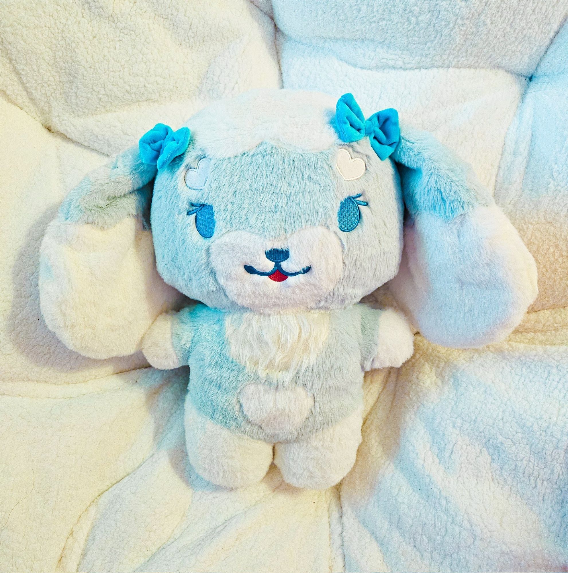 A stylized bipedal fluffy mostly blue with various white accents lop-bunny character plushie sat on a giant white cushion.
The plushie has two blue bows above her two long and floppy ears.