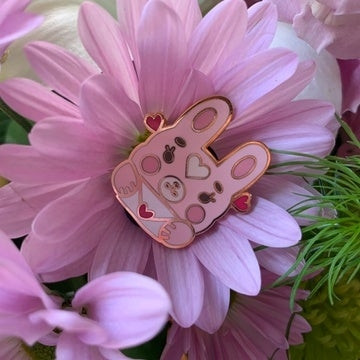 A metal pin on a pink flower. The pin is a stylized chibi bunny character holding a love letter envelope. 