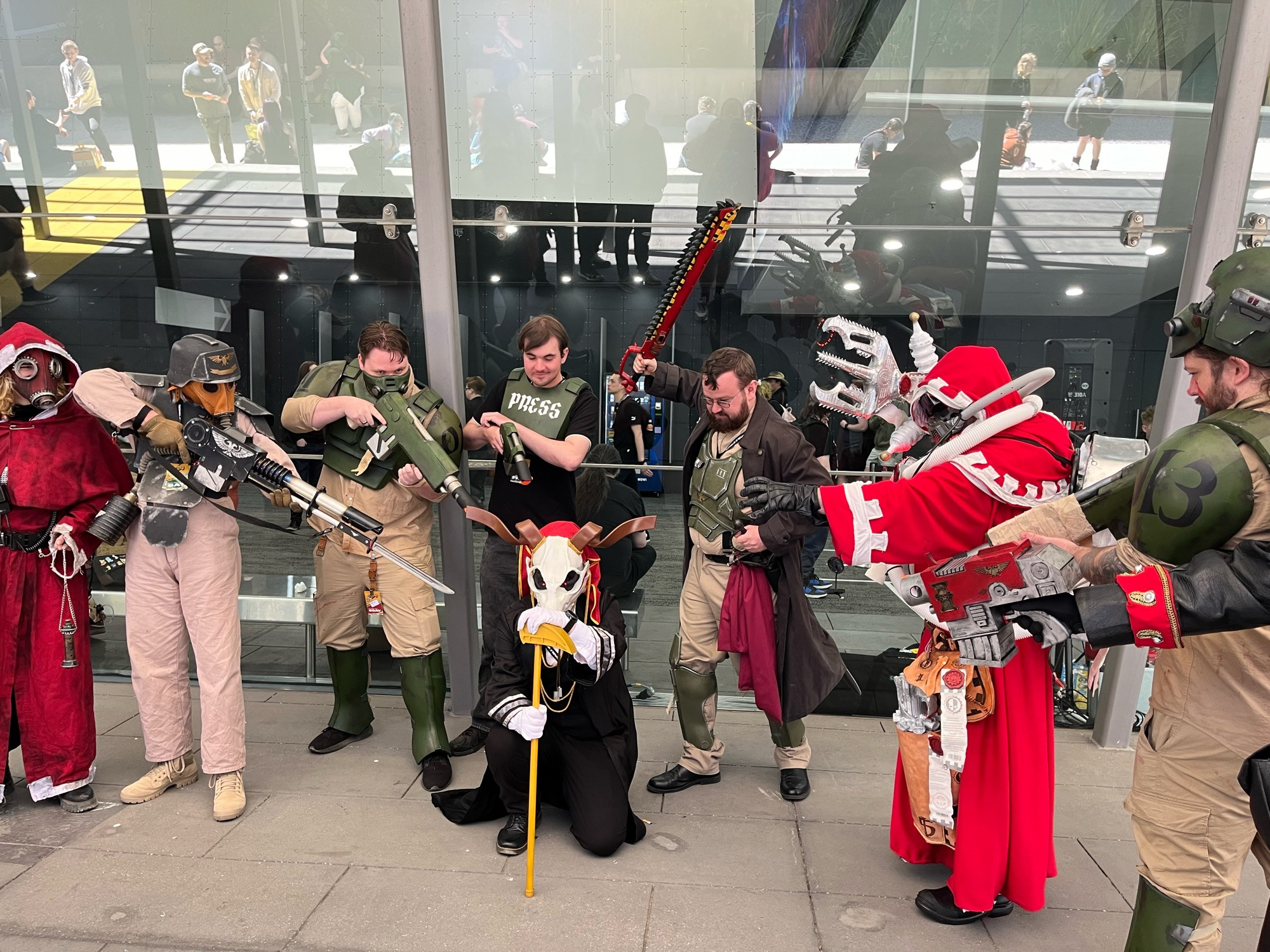 Elias Ainsworth kneeling whilst surrounded by warhammer 40k cosplayers all pointing their weapons at him.