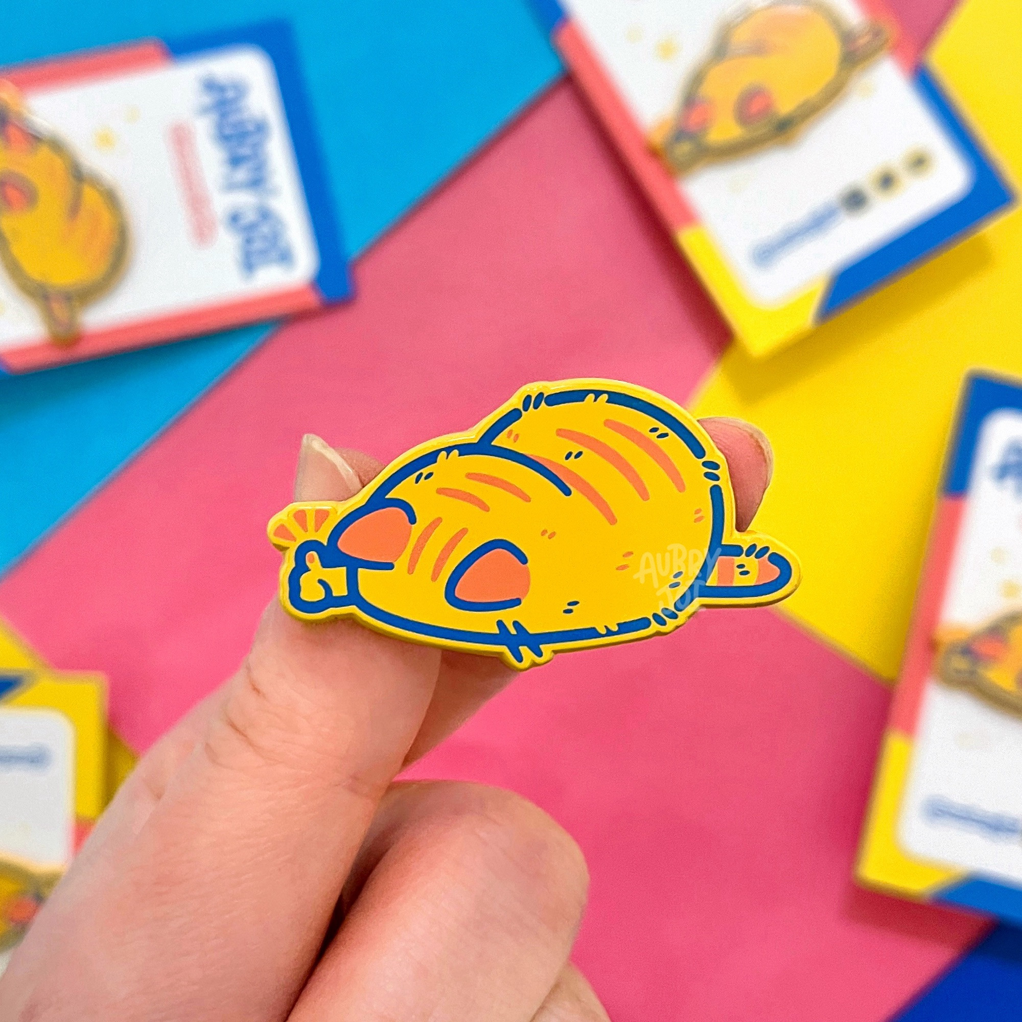 A hand holding an enamel pin of a yellow-orange cat, lying like a loaf, with its face planted down on the ground. It has a paw held out in a thumbs up. The cat has light red ears & tabby stripes, and a blue outline. Some other pins are laying in the background on brightly colored paper.