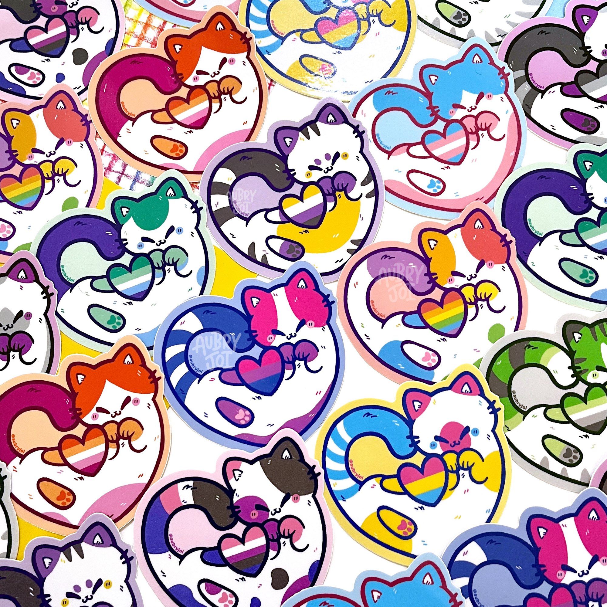 A photo of many overlapping stickers of cats curled around hearts that are striped like various pride flags. The cats’ colors match their flag heart. The flags included are: rainbow, lesbian, bisexual, pansexual, transgender, asexual, nonbinary, genderfluid, aromantic, and gay.