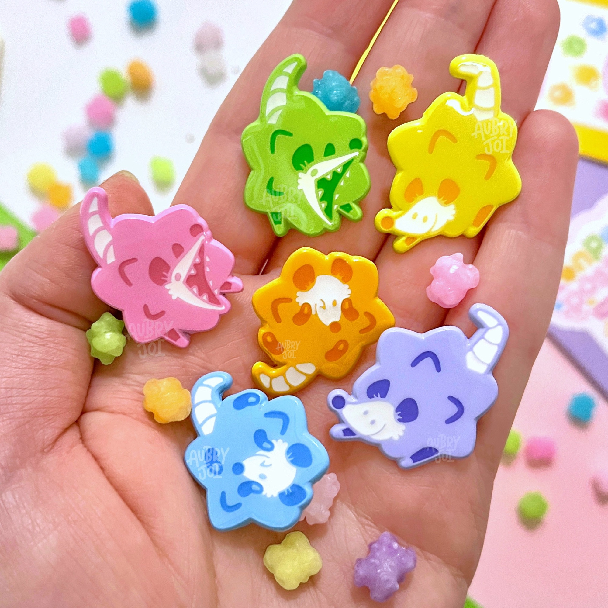 A photo of a hand holding 6 colorful enamel pins. They’re each of an opossum that is made to look like konpeito. They’re pastel, each a color of the rainbow: pink, orange, yellow, green, blue, purple. They look small and shiny. There is some real konpeito scattered amongst them.