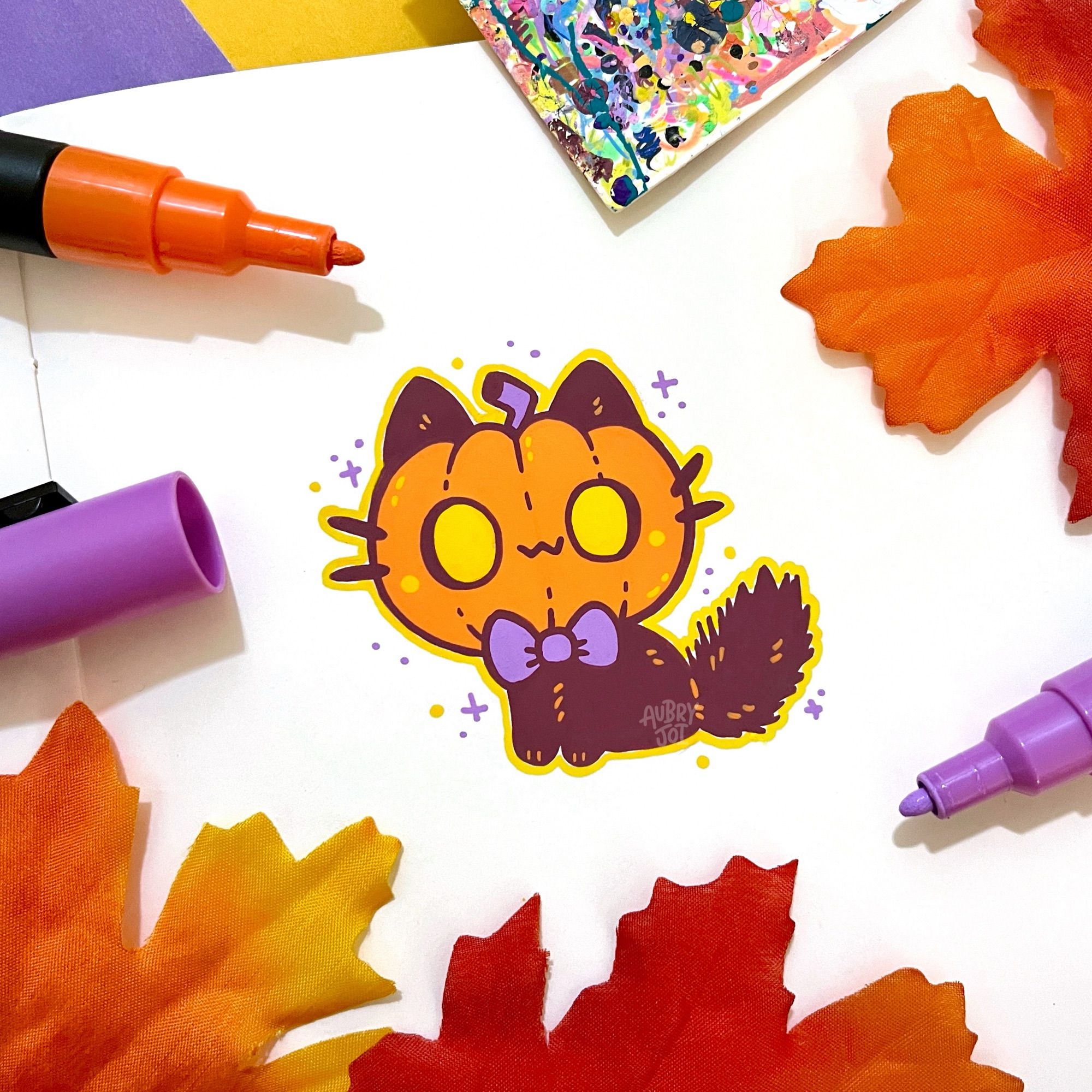 A paint pen illustration in a sketchbook of a small black cat with a jack-o-lantern head and a little purple bow tie. There are small colorful sparkles around them. Fake leaves and paint pens are scattered around the doodle.