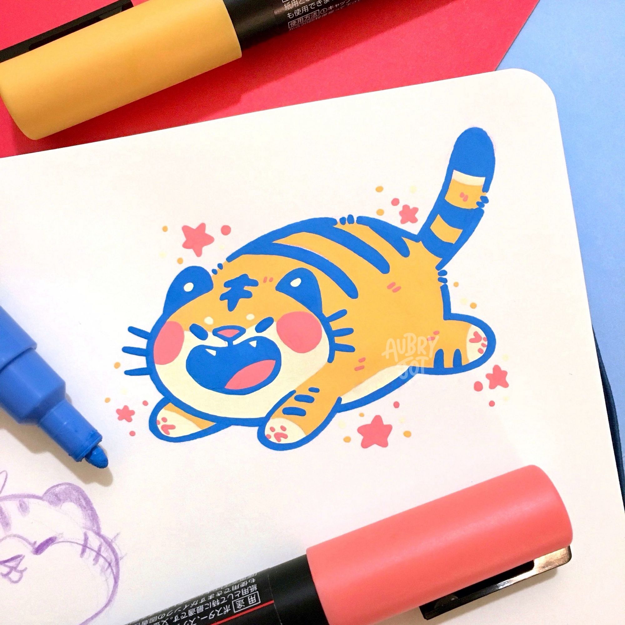 A small paint pen doodle of a round, cute tiger leaping and roaring. It’s colored with bright orange and blue. Its mouth is wide open, fangs showing, and its eyes are closed. It has big pink circular blush marks on its cheeks. Small stars and dots are doodled around it.