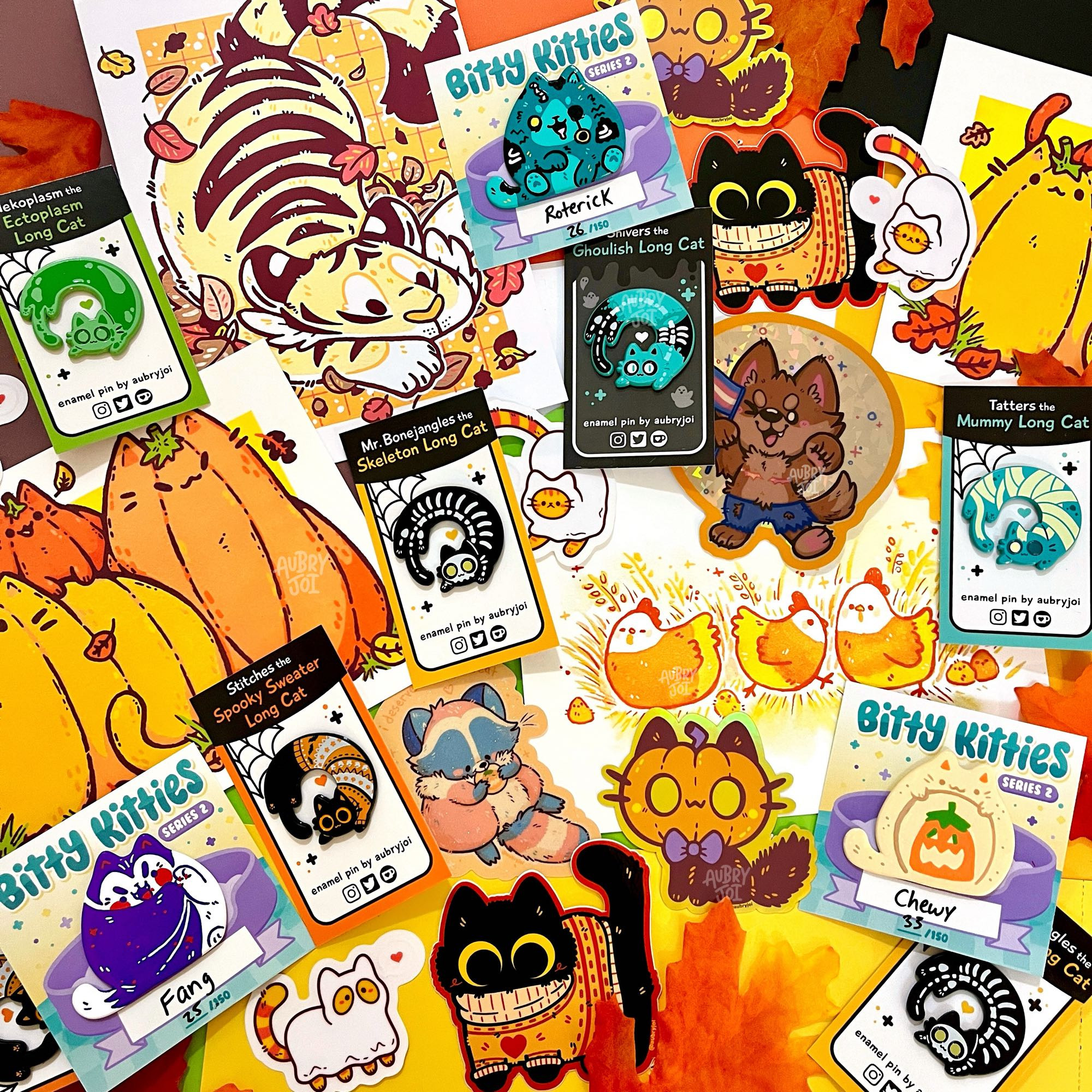 A photo of a large spread of merchandise by Aubry Joi. It’s all fall/Halloween themed, including spooky long cat enamel pins, pumpkin cat & candy corn chicken art prints, trans werewolf & black cat stickers, and more. Everything is very cute and colorful, laying on colorful paper with fake fall leaves scattered around the work.