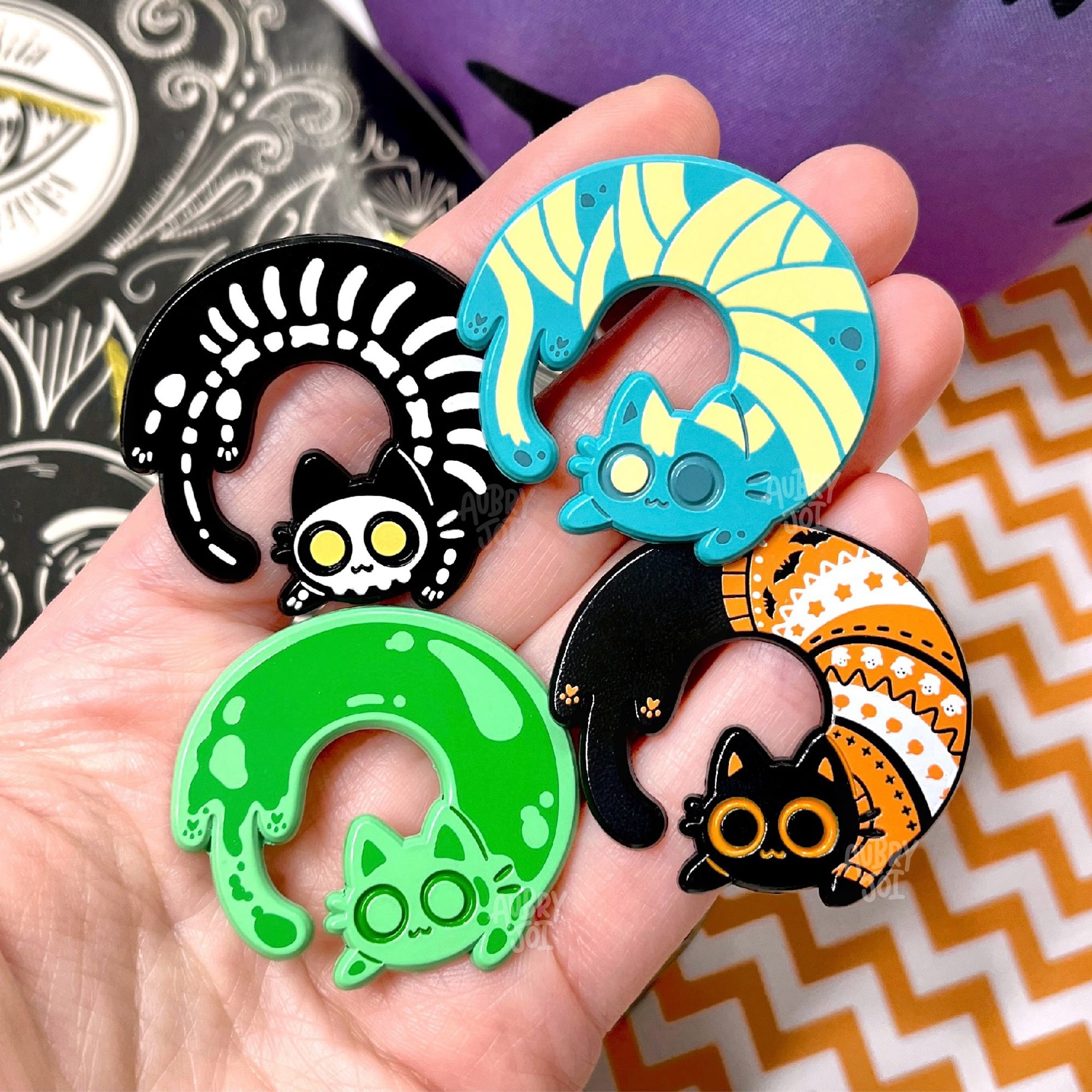 A hand holding four enamel pins of long cat curled into circular shapes. Each cat is themed after something Halloween-related: one is a black cat with a white skeleton printed on it and yellow eyes; one is a green cat with darker green slime/blob shapes on it; one is a dark teal cat with mummy wrappings on it; the last is a black cat with an orange & white Halloween sweater on.