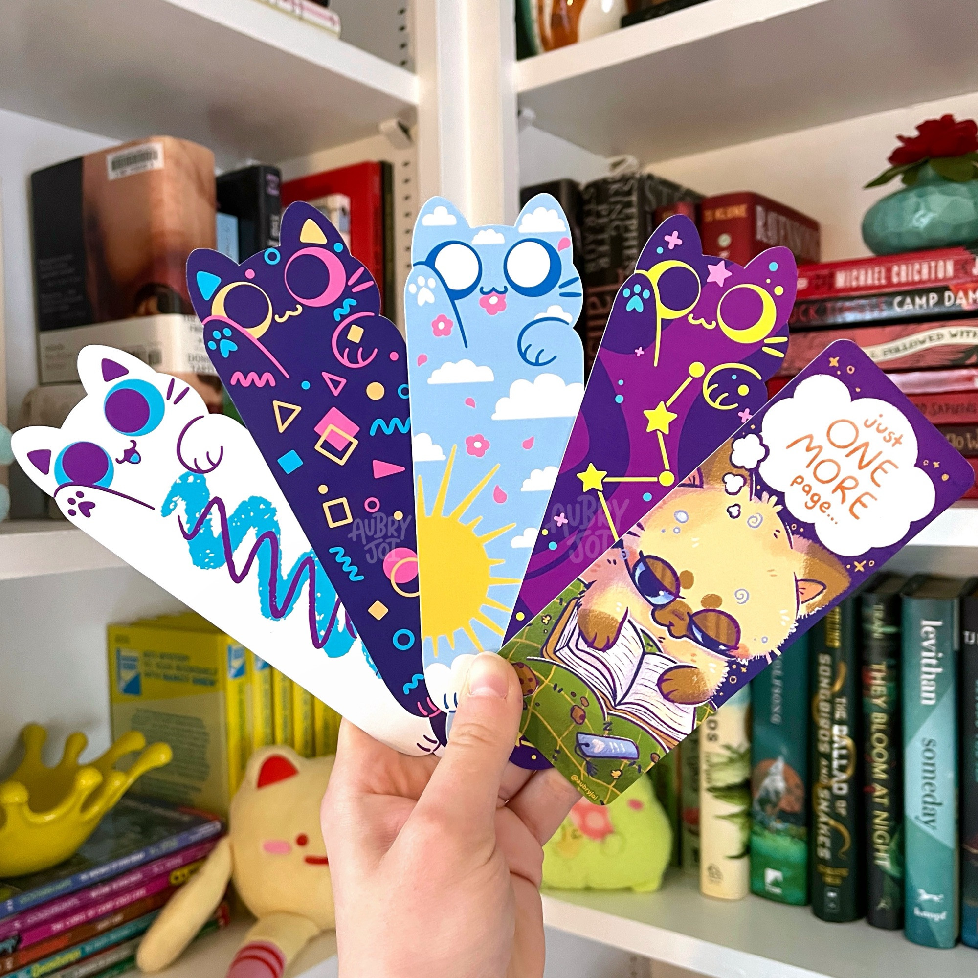 A hand holding 5 diecut bookmarks fanned in front of a bookshelf. They are, in order: green alien long cat; retro cup long cat; retro arcade carpet long cat; sunny sky long cat; purple constellation long cat; a sleepy siamese cat reading a book in bed, thought bubble over his head reading “just one more page…”
