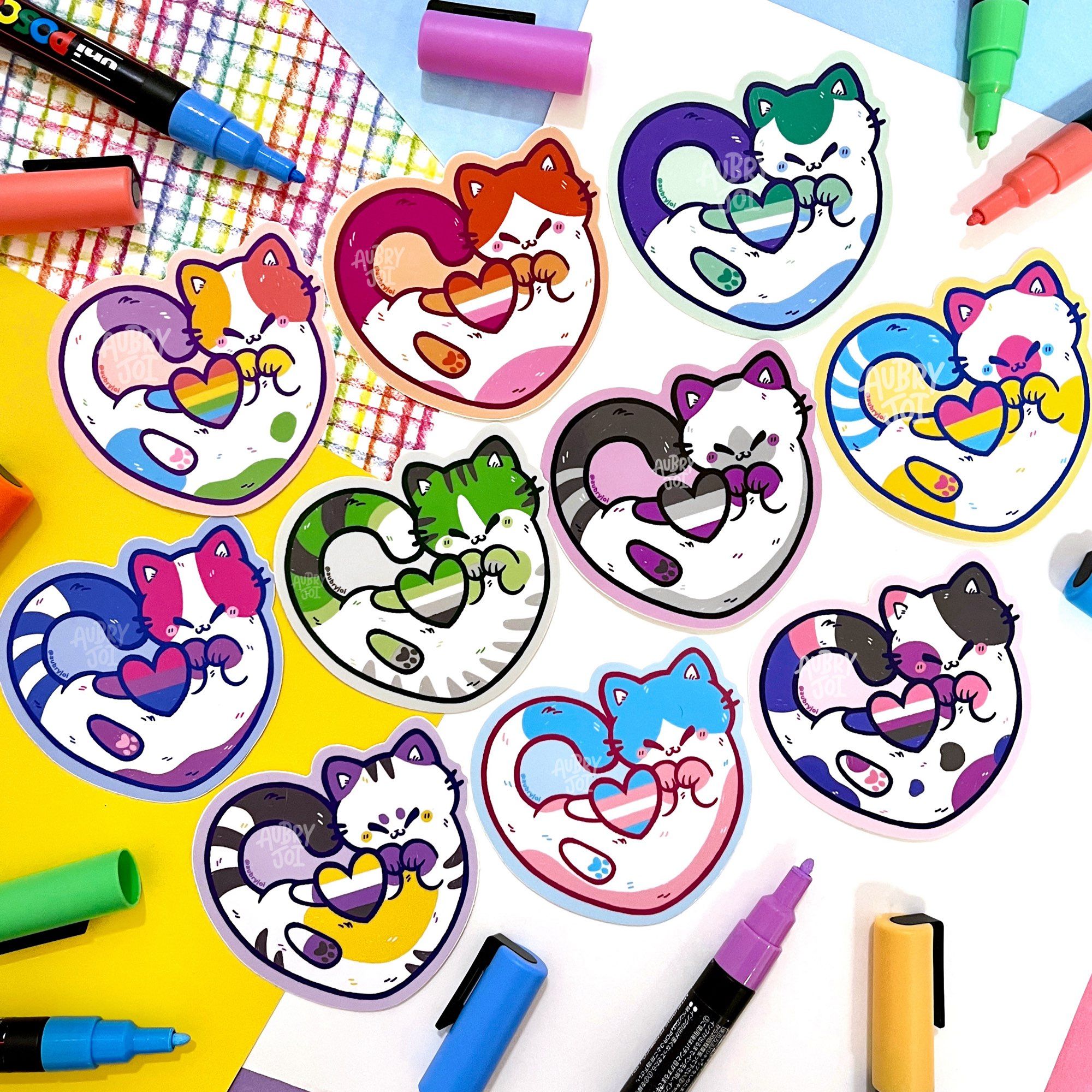 A photo of 10 stickers of cats curled around hearts that are striped like various pride flags. The cats’ colors match their flag heart. The flags included are: rainbow, lesbian, bisexual, pansexual, transgender, asexual, nonbinary, genderfluid, aromantic, and gay.