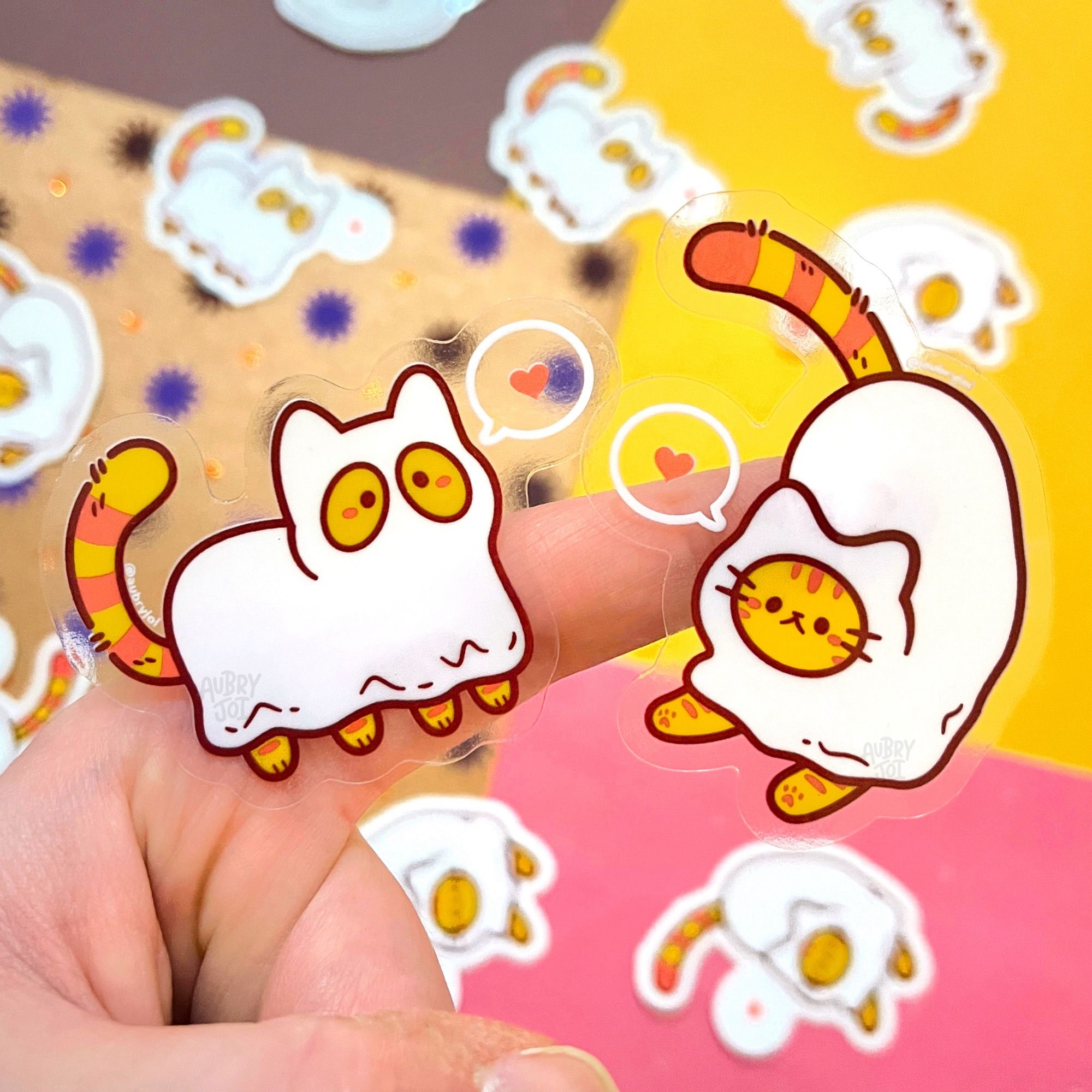 Two stickers stuck to a finger; each sticker is of a cute ghost cat. The one on the left is standing on all four paws, and has a sheet over its while body. An orange tabby tail is poking out, and two eyes on an orange face are visible. The one on the right is jumping downward, and has a circle cut in its sheet so its whole orange tabby face as show through. Both ghosts have white speech bubbles with red hearts in them. The stickers are printed with transparent borders.