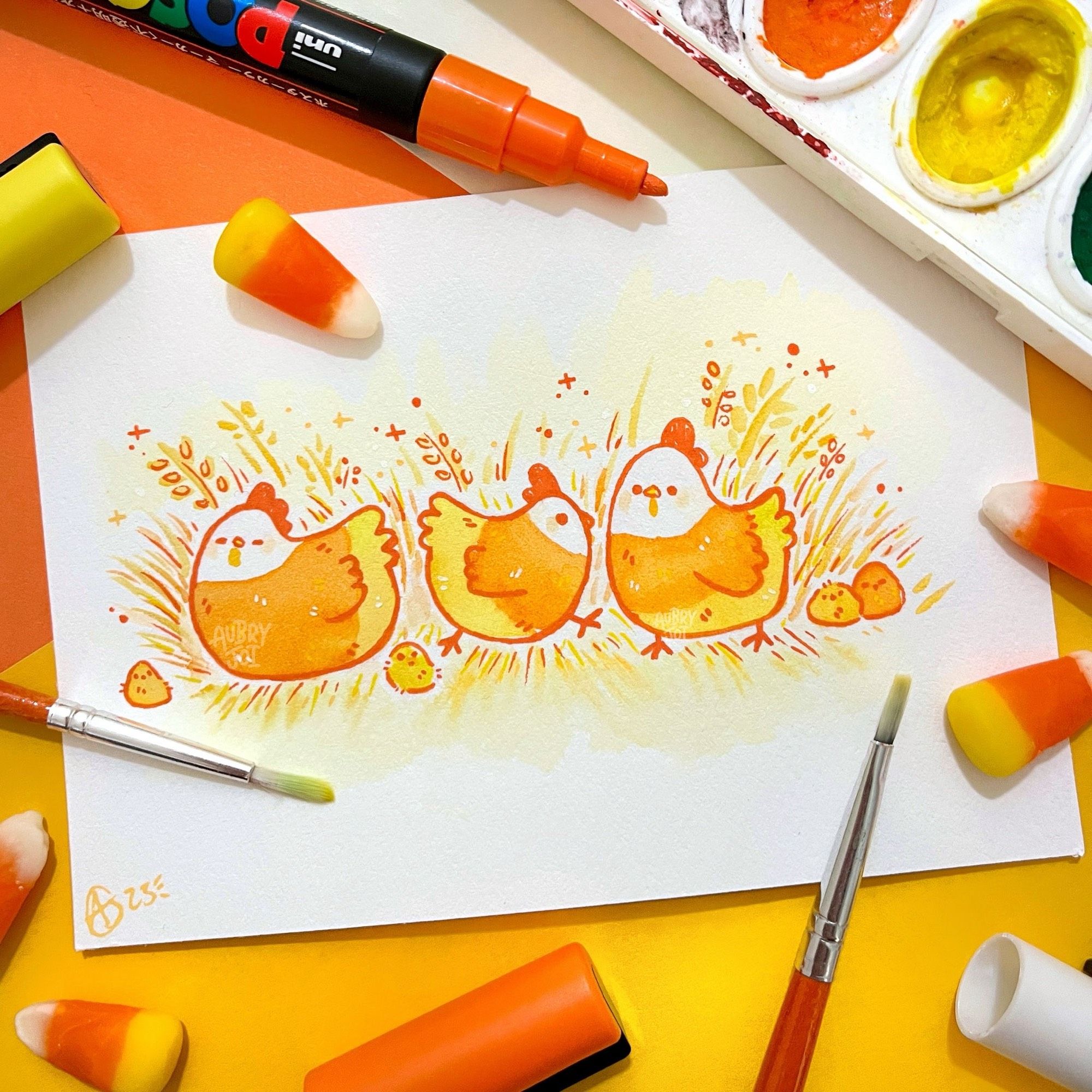 A watercolor painting of three cute chickens. Each is colored to resemble candy corn: white heads, orange middles/wings, and yellow bottoms. There are several round chicks amongst them, in various shades of orange. They are all sitting in some orange & yellow grass, with small sparkles around them. The painting is laid among some paint brushes & Posca paint pens, with a few pieces of real candy corn scattered around.