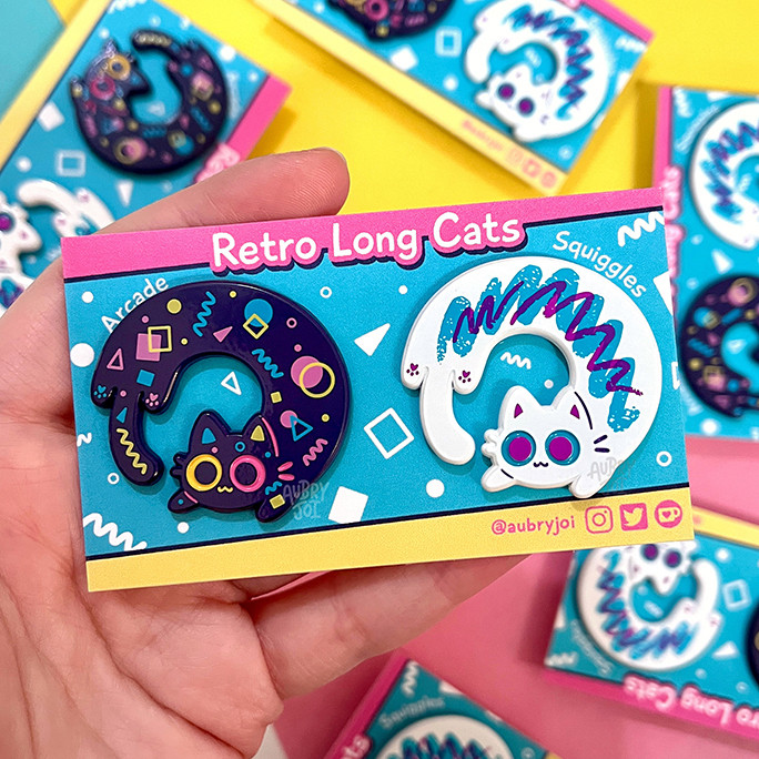 My hand holding two enamel pins of long, circular cats. The one on the left is dark purple with a colorful arcade-carpet pattern on it of geometric shapes & lines. The pin on the right is white with the iconic “jazz cup” design, a teal squiggle with a purple squiggle on top. The pins are on a backer card that shows their names & the title of the set: Retro Long Cats, named Arcade & Squiggles.