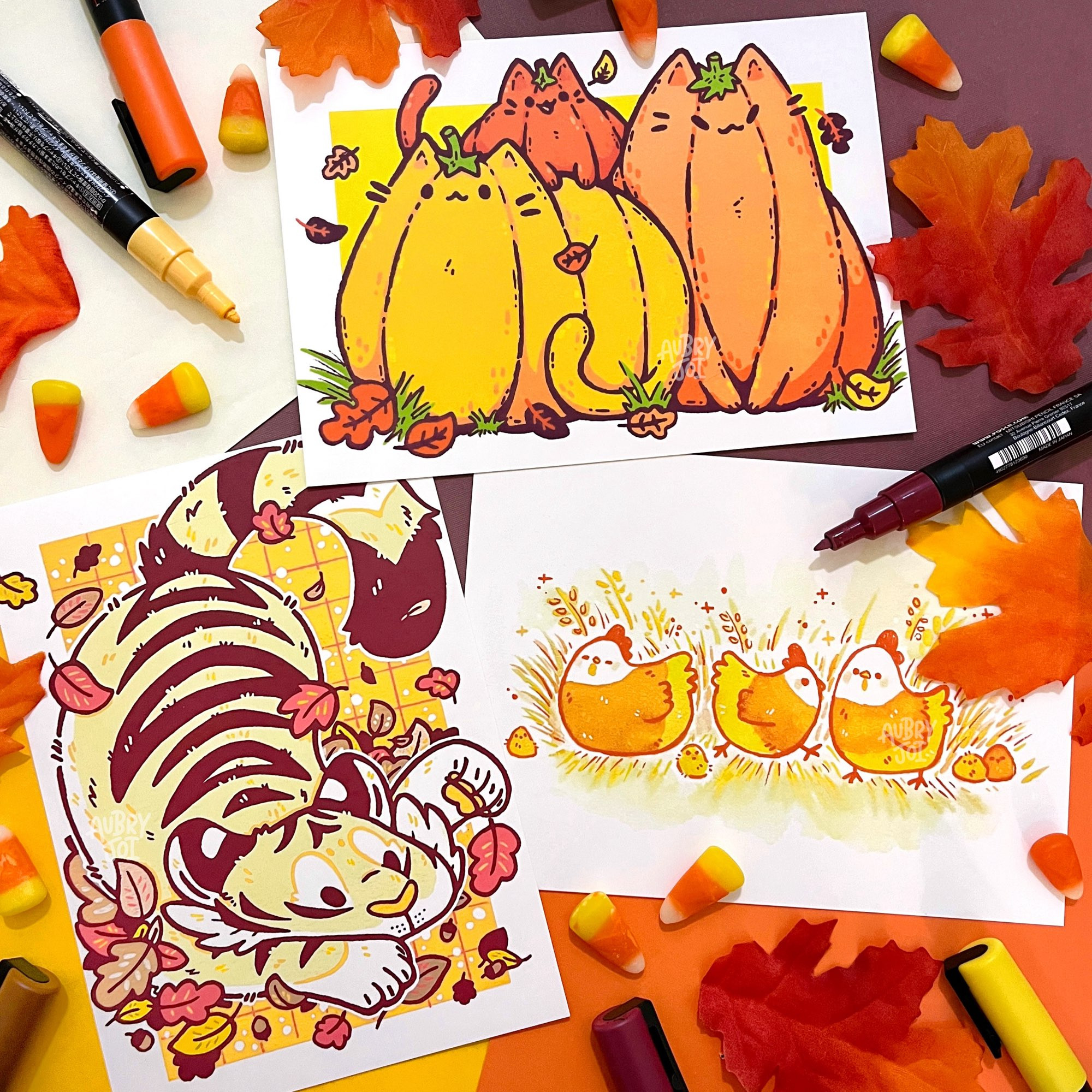 A photo of three art prints, surrounded by fake autumn leaves, candy corn, & paint pens. One is of three pumpkin cats. One is of a white tiger with dark red stripes, playing in a pile of fall leaves. The last print is of three chickens colored like candy corn, painted with watercolors.